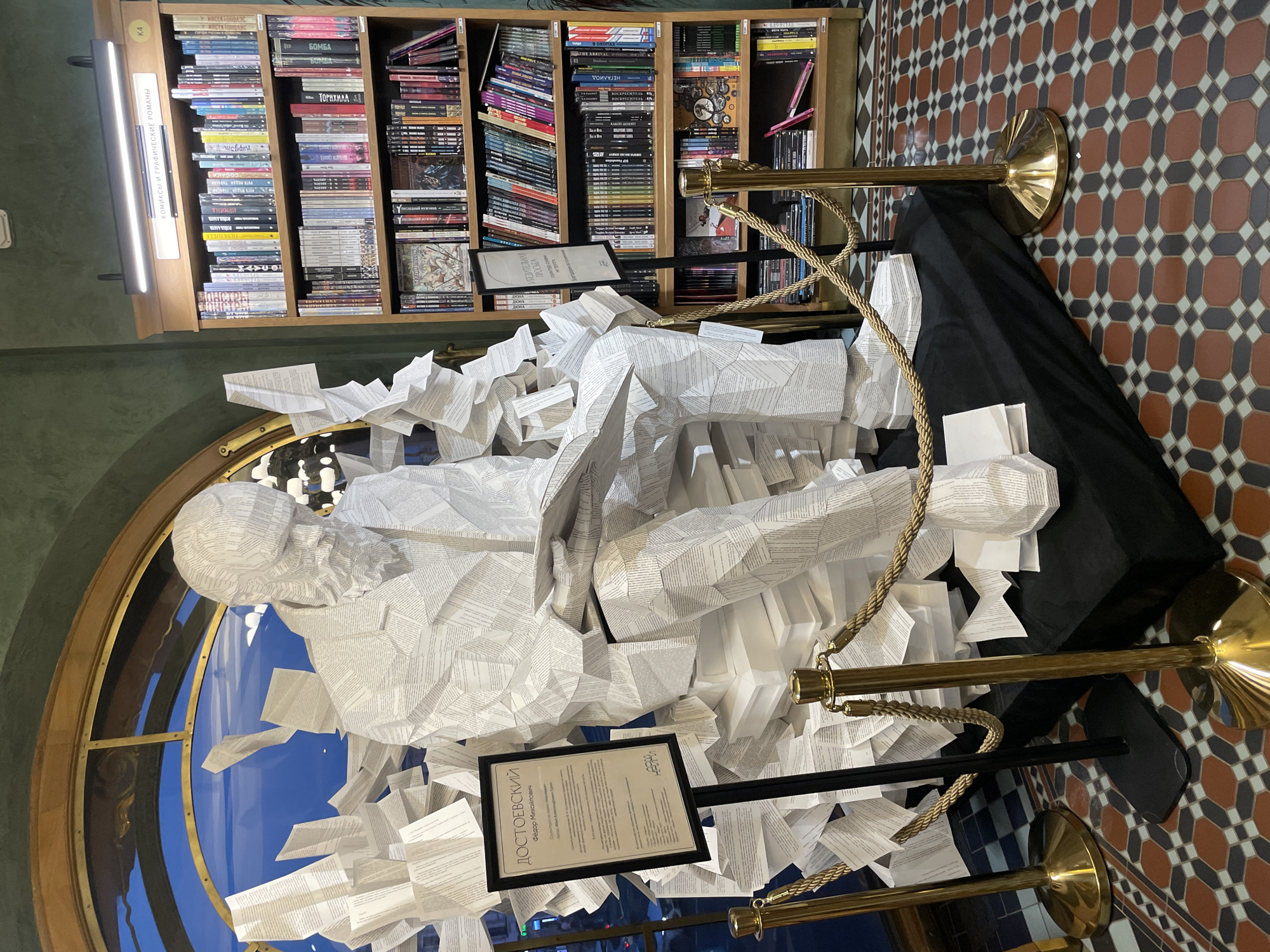 A large, multi-faceted sculpture of Fyodor Dostoevsky, constructed from pages of text, is centrally displayed within the Dom Knigi (Singer House) bookstore on Nevsky Prospekt in Saint Petersburg, Russia. The statue depicts Dostoevsky seated, wearing a suit and beard, looking down at an open book in his hands. It rests on a black, fabric-draped platform, surrounded by numerous loose and folded paper forms that extend upwards and outwards, creating an impression of scattered pages or an erupting literary work.
To the left of the sculpture, a black framed sign on a stand identifies the subject as "ДОСТОЕВСКИЙ Федор Михайлович" (Dostoevsky Fyodor Mikhailovich) and credits the work to artist Ilya Aleksandrovich Bange as part of the "Faces of Russian Literature" series. To the right, another framed sign on a stand displays "УБЕДИТЕЛЬНАЯ ПРОСЬБА" (Earnest Request). Gold-colored stanchions with braided gold ropes delineate a viewing area around the sculpture.
In the background to the left, a large, ornate arched window with a gold-colored frame reveals a bright blue sky and distant architectural details outside. To the right of the sculpture, a tall wooden bookshelf is fully stocked with various books. Above the top shelf, a sign reads "КОМИКСЫ И ГРАФИЧЕСКИЕ РОМАНЫ" (Comics and Graphic Novels), illuminated by a horizontal LED light fixture. The walls surrounding the display are a dark, textured green. The floor features a distinct geometric pattern of octagonal and square tiles in shades of terracotta, dark brown, light gray, and dark gray.