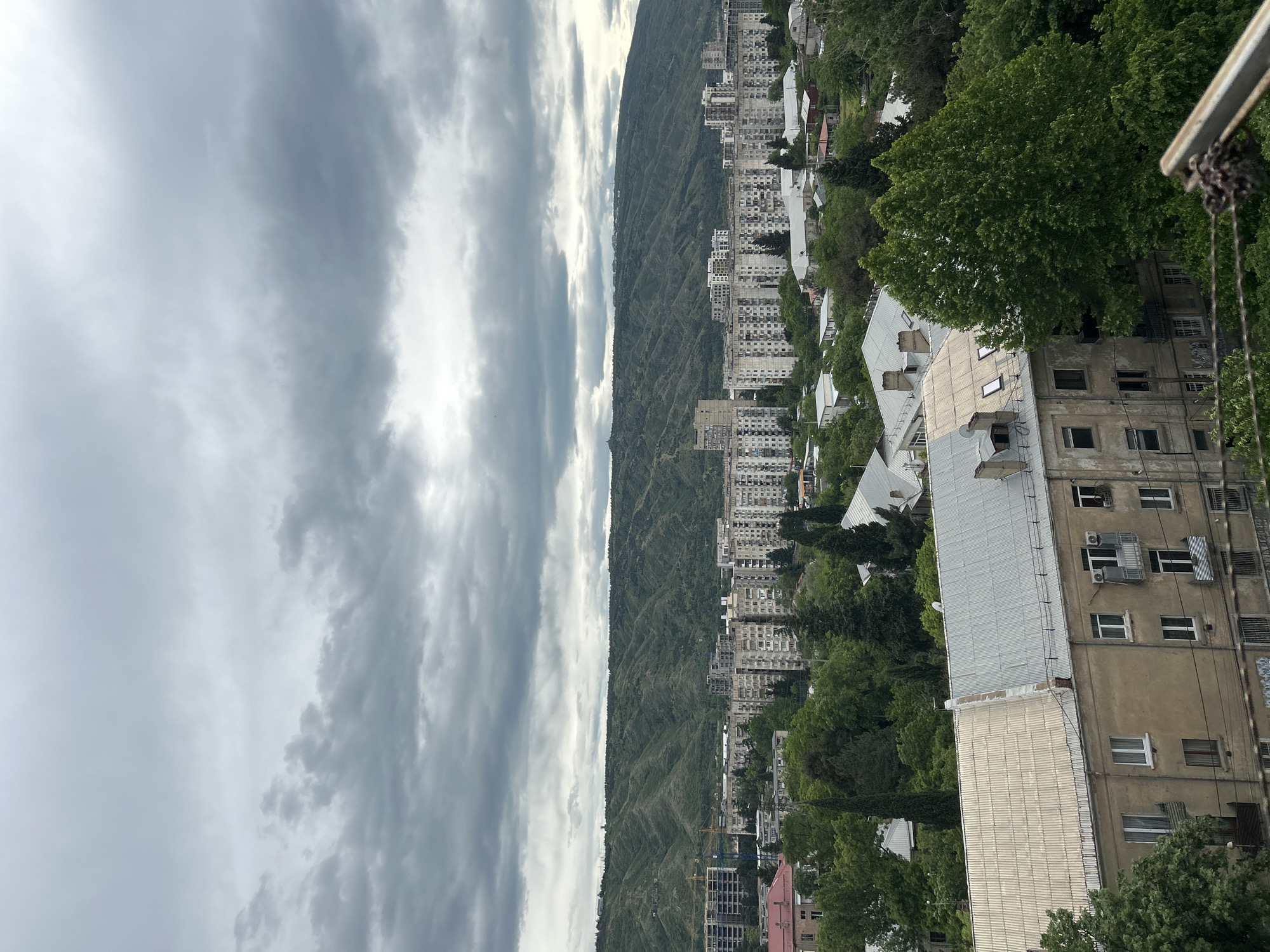 The photograph depicts an elevated cityscape with a backdrop of a tree-covered mountain ridge. The sky is overcast, filled with gray clouds. In the middle ground, dense residential buildings are mixed among the trees, forming an urban landscape. The photograph seems to have been taken on the edge of the city, showcasing the natural border with the mountains. It is likely a view from Tbilisi, Georgia.