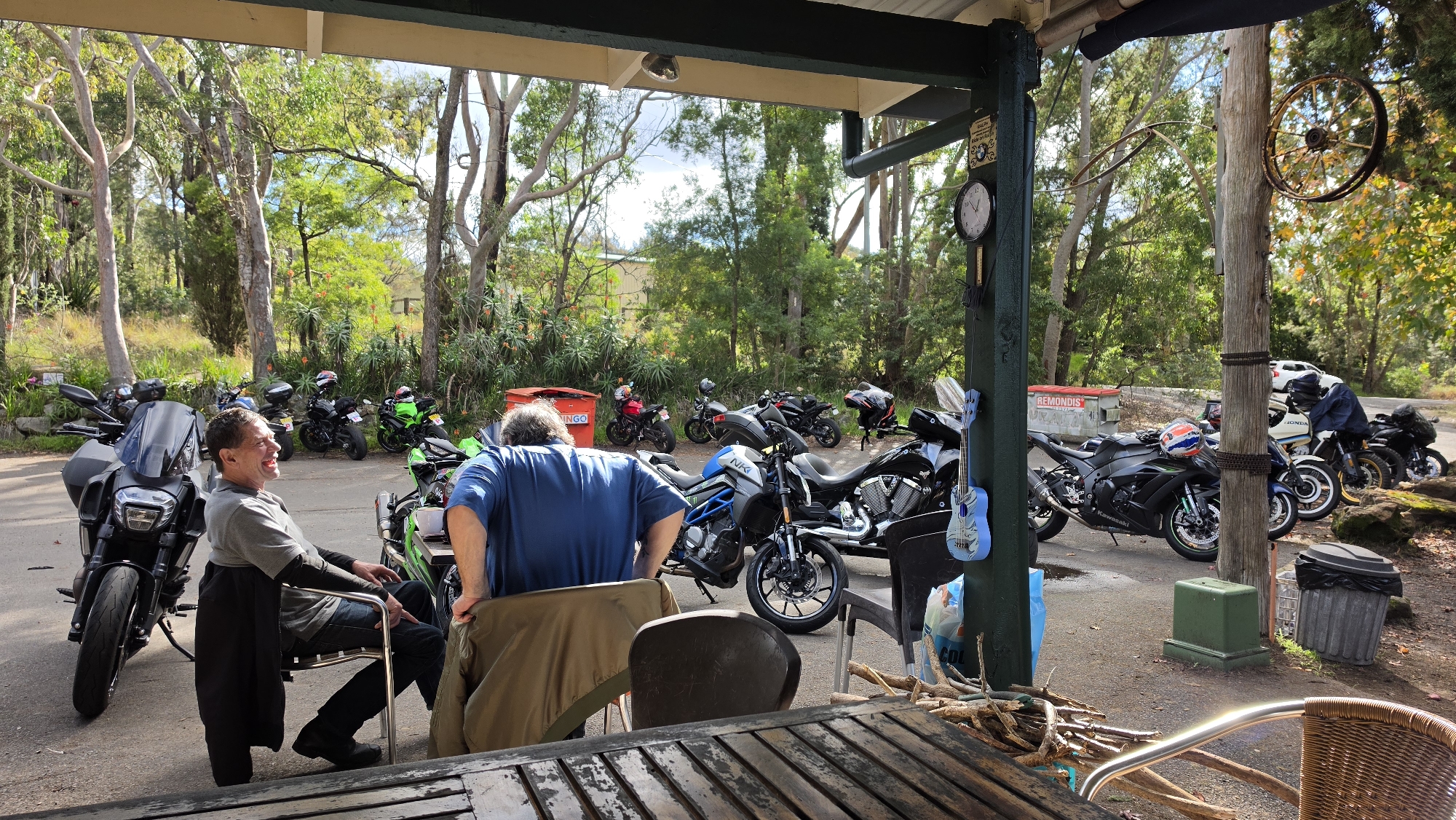 The image captures a sunny outdoor scene at a roadside establishment in Mount White, Australia, likely a cafe or biker-friendly stop, situated within a wooded environment. In the foreground, a wooden table with visible wood grain and metal railings suggests an outdoor seating area. Two men are present. One man, seated on a metal chair with a black jacket draped over it, faces left and appears to be laughing. He is wearing a grey long-sleeved shirt and dark trousers. The second man, wearing a blue t-shirt, stands with his back mostly to the camera, facing a row of motorcycles.
A collection of motorcycles is parked behind the men. From left to right, there is a large, dark motorcycle with a prominent windscreen, followed by several other motorcycles of various colors, including green, black, and a blue and grey model. Some of these motorcycles have helmets on them, and a blue decorative guitar is hanging on a green pole near the foreground.
In the background, a vibrant green wooded area with tall trees dominates the landscape. There are glimpses of other structures, including a red bin with "REMUNDIS" written on it, and what appears to be a clock on a wooden post. The lighting indicates a bright, daytime setting. The overall atmosphere suggests a gathering of motorcycle enthusiasts taking a break in a natural, semi-rural location.