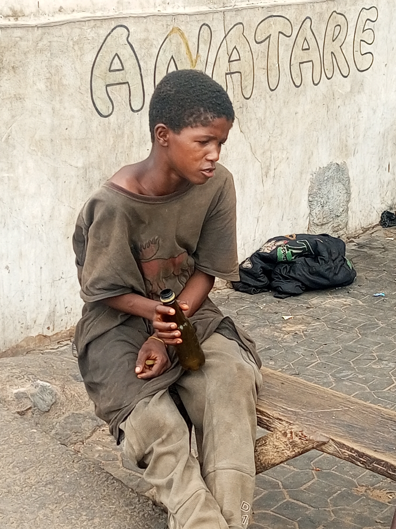 The image depicts a young individual, appearing to be male, seated outdoors against a light-colored, textured concrete wall in the city of Unknown, Unknown. The individual has a dark complexion and short, dark, tightly curled hair. They are dressed in an oversized, dark olive-brown long-sleeved top and light brown, possibly tan, trousers; the left trouser leg displays a faint "D 1" marking near the ankle. The individual holds an uncapped dark glass bottle, which appears to be brown or dark green, with a yellow-gold metallic rim in their right hand. A small, light-colored object is held in their left hand. Their gaze is directed downwards and to the right, with the mouth slightly agape. They are seated on a simple, weathered wooden bench, which extends from the bottom right. The wall behind features large, black-outlined block letters spelling "ANATARE," with some letters exhibiting faded yellow infill. The ground is paved with dark grey interlocking pavers. To the right of the individual, a crumpled black item, possibly a jacket or bag with green and orange design elements, rests on the ground amidst scattered small pieces of debris.
