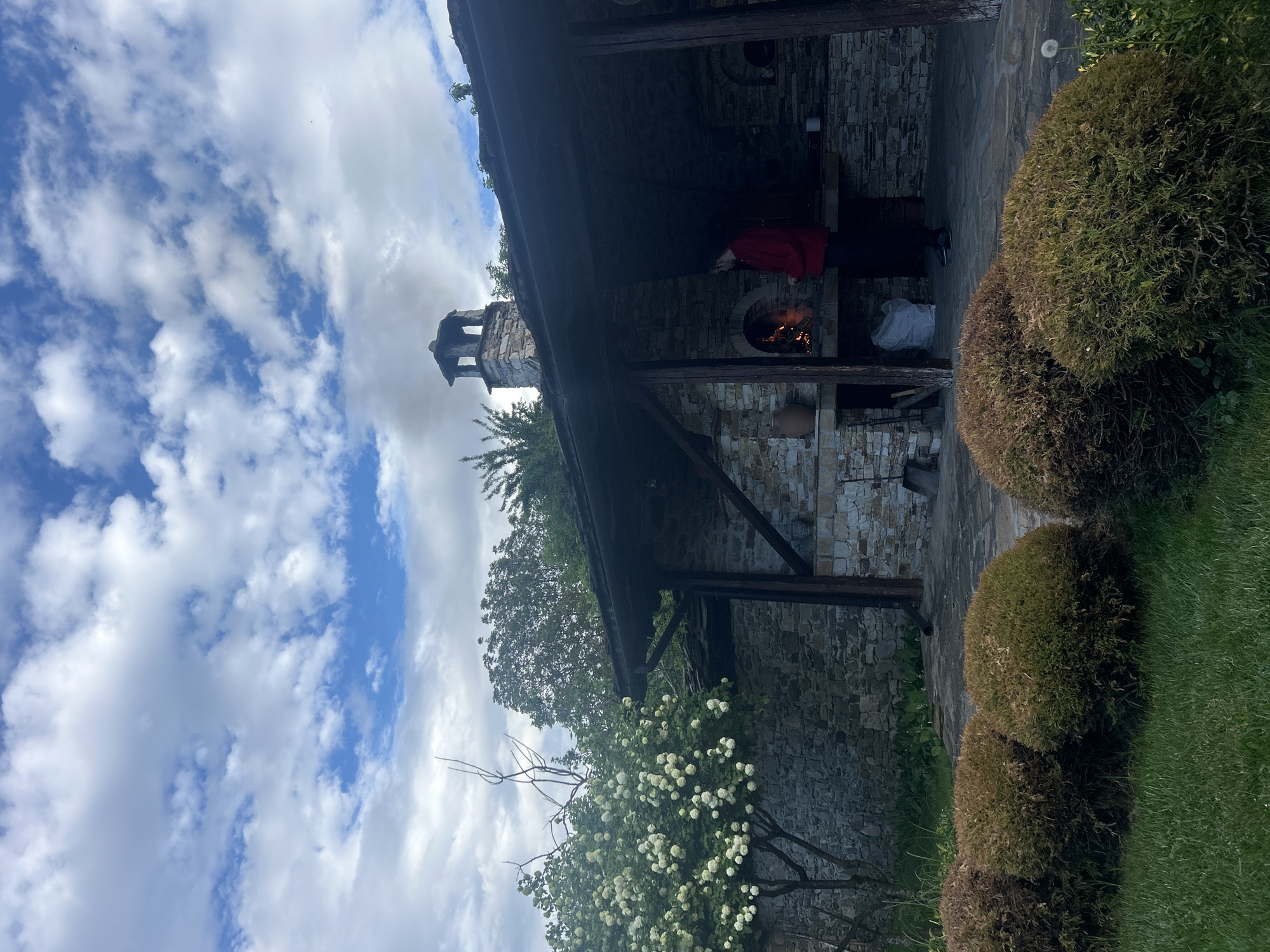 The scene depicts an outdoor stone structure with a chimney, likely a grill or oven, located in Dolen, Bulgaria. A person wearing a red garment is near the oven, where a fire is burning. The structure is partially covered by a dark, overhanging roof. The sky is visible, exhibiting a mix of blue sky and scattered clouds. Vegetation, including flowering bushes and rounded shrubs, surrounds the structure. The area appears to be a well-maintained garden.