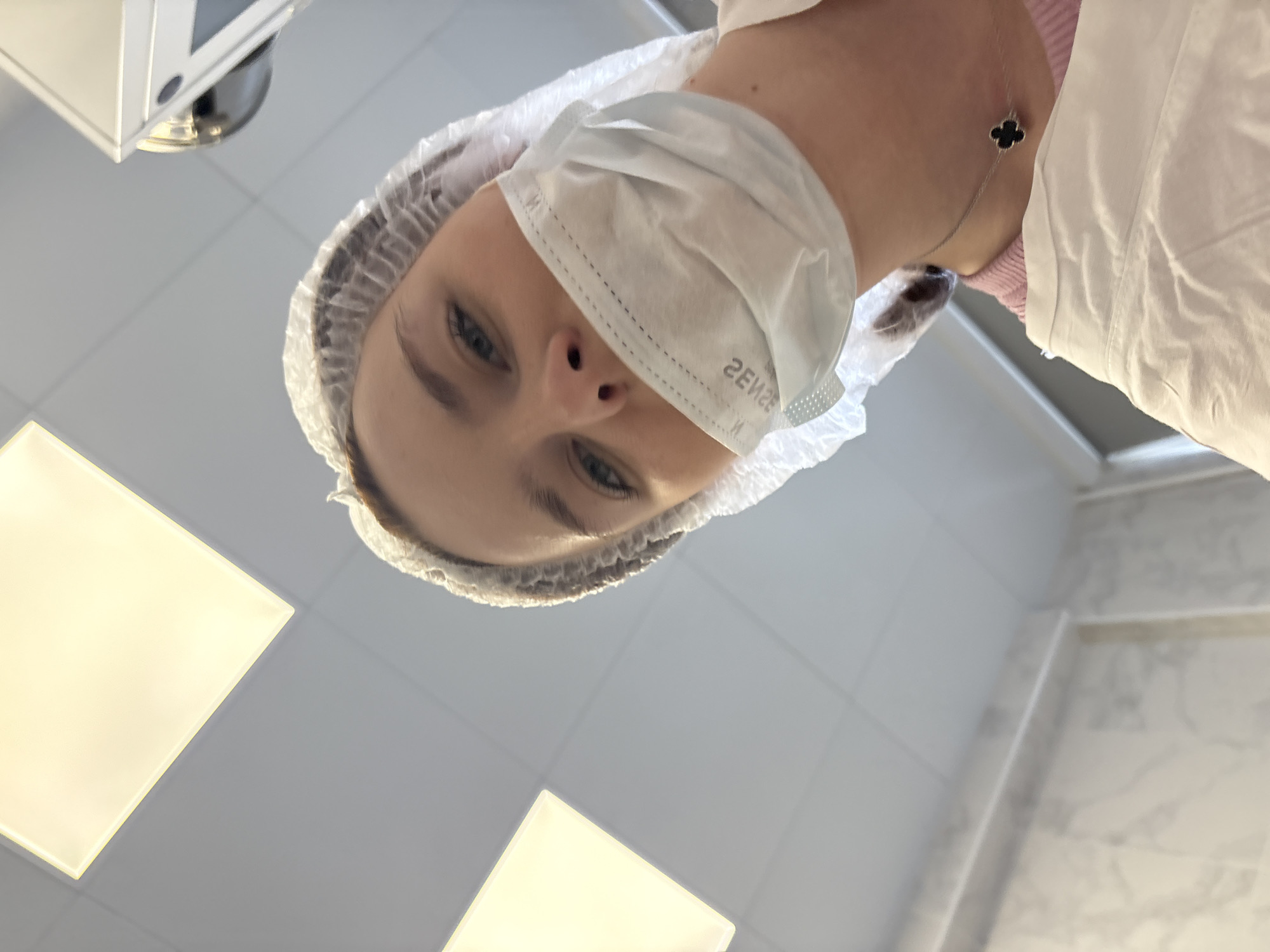 The image shows a person from a low angle, likely a patient's perspective. The person is wearing a surgical cap and a white face mask, suggesting a medical setting. Their eyes are visible, and they appear to be looking down at the camera. A necklace with a dark, flower-like pendant is visible. The background consists of white tiles and a fluorescent ceiling light. The location cannot be determined definitively from the image, so the information is not confirmed to be in Saint Petersburg, Russia.