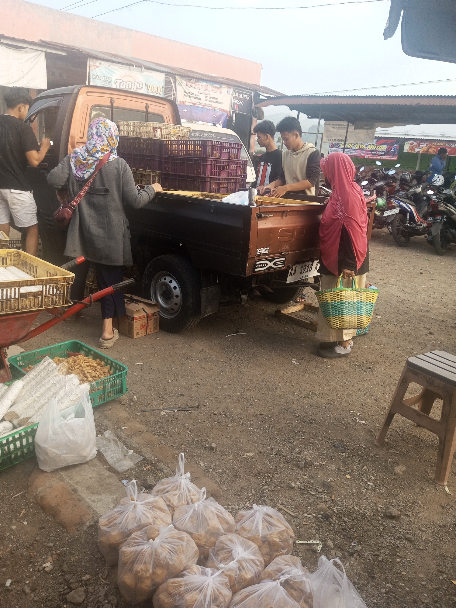 The image depicts a bustling outdoor market scene, likely in Kalibeber, Indonesia, at what appears to be dawn or dusk, given the soft, diffused lighting. A small orange utility truck is central to the frame, its bed filled with purple crates. Several individuals are engaged in activities related to the truck and its contents.
In the foreground, on the left, a person in a grey jacket and floral headscarf is leaning against the truck bed. To their left, a wheelbarrow with a yellow crate containing white, cylindrical items and another green crate filled with peanuts or similar small items are visible. In the lower right of the frame, several large plastic bags filled with what appear to be potatoes or root vegetables are scattered on the ground. A small wooden stool stands to the far right.
Behind the truck, two young men are interacting near the driver's side. One, wearing a black t-shirt, is partially obscured by the truck. The other, in a beige hooded sweatshirt, is looking down, seemingly engaged with something on the truck.
To the right of the truck, a person wearing a pink headscarf and a dark outfit is holding a woven basket and looking towards the ground. Several motorcycles are parked behind them, suggesting a public thoroughfare or a dedicated parking area within the market.
The ground is composed of dirt and gravel. In the background, market stalls with awnings and signage can be seen, indicating a commercial area. Banners with text, some in Indonesian, are displayed above the stalls. The overall atmosphere is one of active commerce and daily life in an Indonesian market.