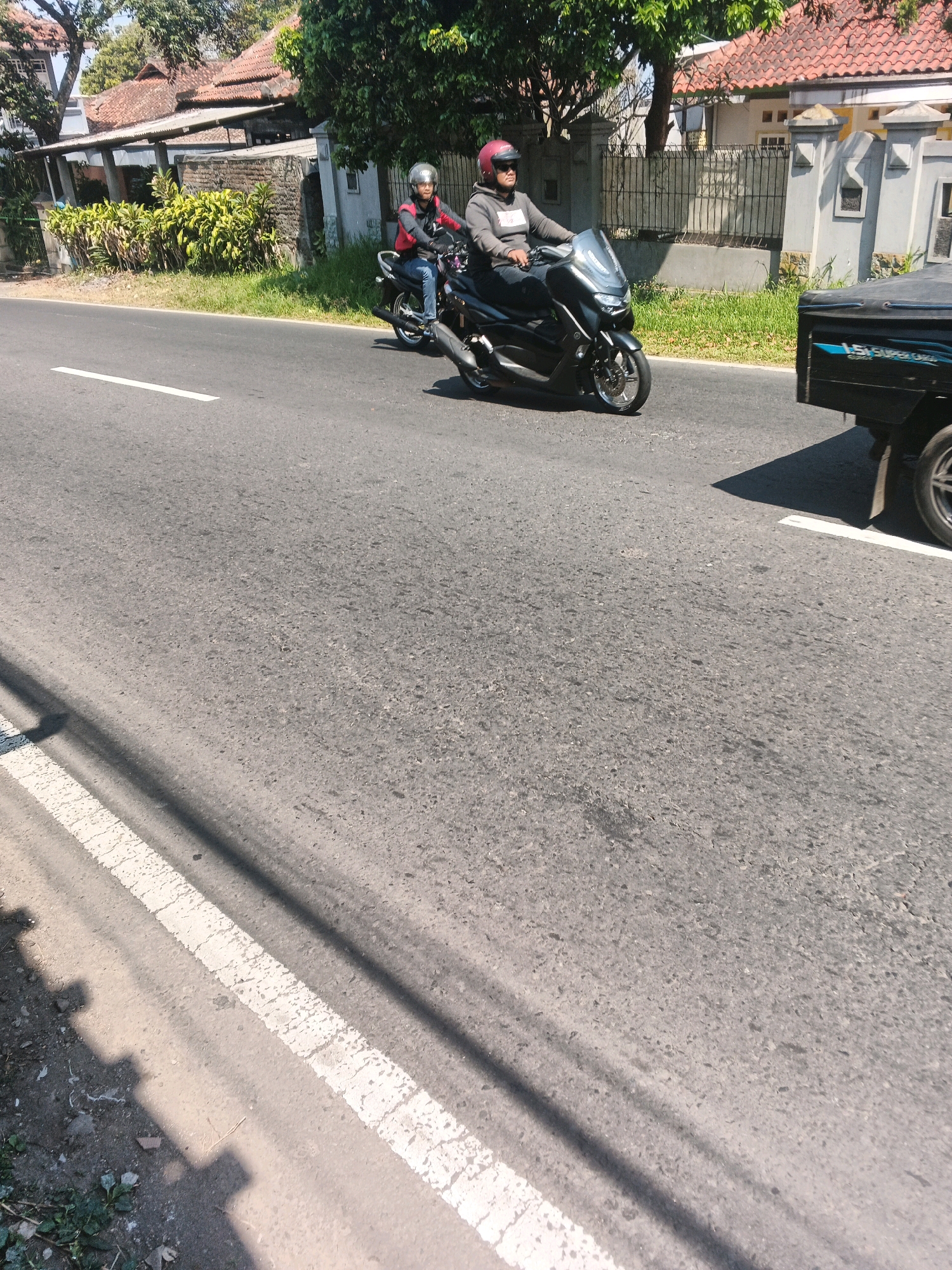 In Bandungan, Indonesia, on a sunny day, two individuals are seen riding a motorcycle. The person in the front is wearing a grey hoodie and a red helmet, while the person behind them is wearing a grey jacket and a red helmet. They are traveling on a paved road with a white line marking the lane. To the right of the motorcycle, a black truck is partially visible, also moving along the road. The background features a residential area with houses, a fence, and lush green trees. The scene captures a moment of daily life on a street in Bandungan.