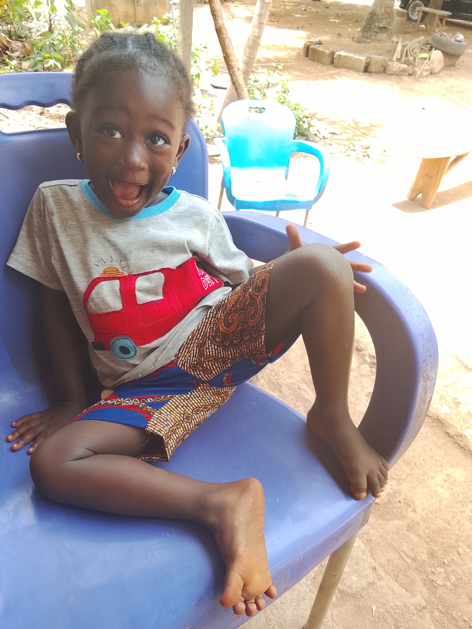 A young Black child with short, dark hair sits on a blue plastic chair outdoors in Akwanga, Nigeria. The child wears a light gray t-shirt with a red fire truck appliqué and patterned shorts. The child's mouth is open in a wide expression. A second blue plastic chair is visible in the background, along with a wooden table. The ground appears to be dirt or packed earth.