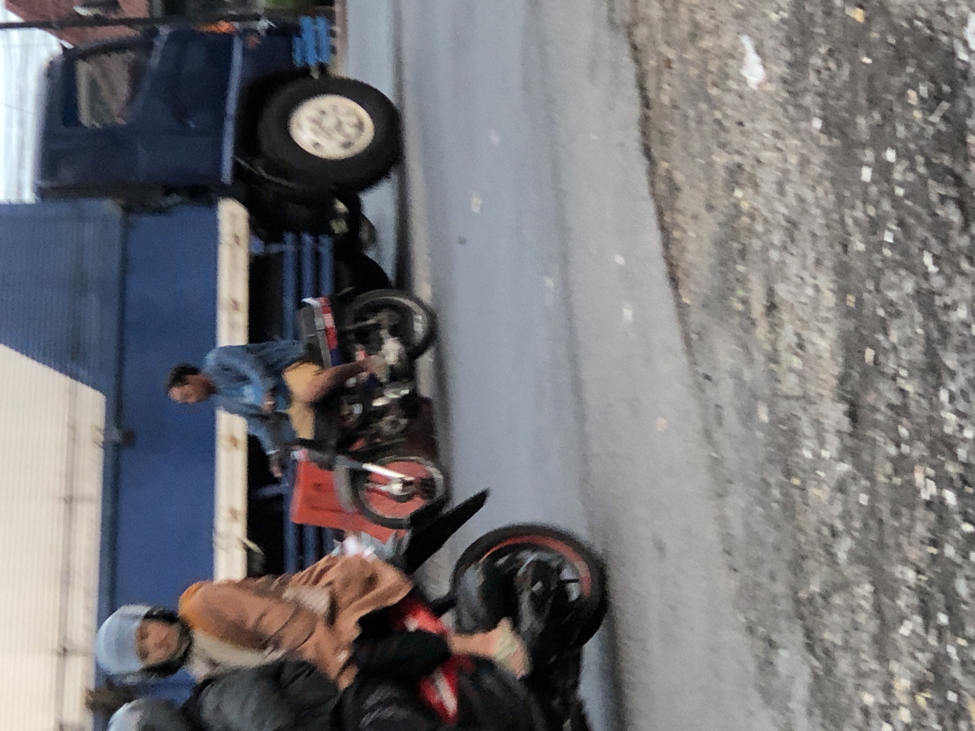 A mid-shot photograph, taken from a slightly elevated angle, captures a street scene in Kludan, Indonesia. The foreground is dominated by asphalt, with a scattering of small stones and debris. In the mid-ground, two motorcycles are visible. The motorcycle on the left is partially obscured by the motorcycle on the right, and its front wheel is in focus, revealing a red rim. A person wearing a light-colored helmet and a tan-colored garment is seated on this motorcycle. Behind this, the second motorcycle is more fully visible, with a rider wearing a blue denim jacket and beige shorts. This motorcycle features a red fender and a silver exhaust pipe. To the right of the motorcycles, a large dark blue truck is parked, its massive rear tire, with a silver rim, prominent in the frame. The truck is positioned on the road, and its side is marked with a white stripe. The background is blurred, showing a light-colored structure and a hint of greenery. The overall impression is that of a busy street with vehicles in motion.