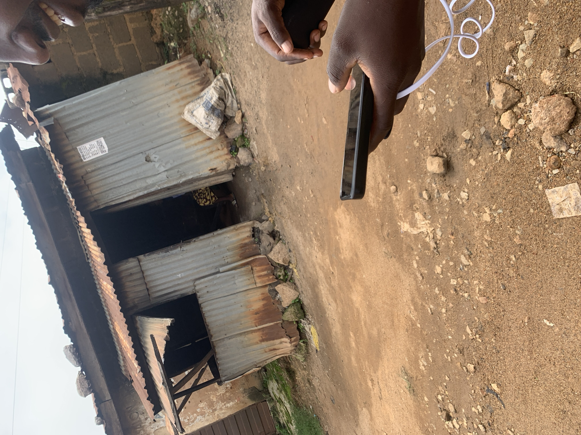 A close-up shot captures a person's hands holding a smartphone with a black case and a white wired headset looped around the fingers. The phone is being held in the foreground, with the screen potentially in use, though it is not clearly visible. In the background, a weathered, corrugated metal structure, likely a stall or small shop, stands on a dirt ground with scattered rocks and patches of green vegetation. The structure appears aged, with rust and some structural damage visible. A large, light-colored bag rests near the entrance of the structure. The overall scene suggests an outdoor urban environment, potentially in Jos, Nigeria, given the architectural style and the type of structures often found in such areas. The lighting indicates it is daytime.