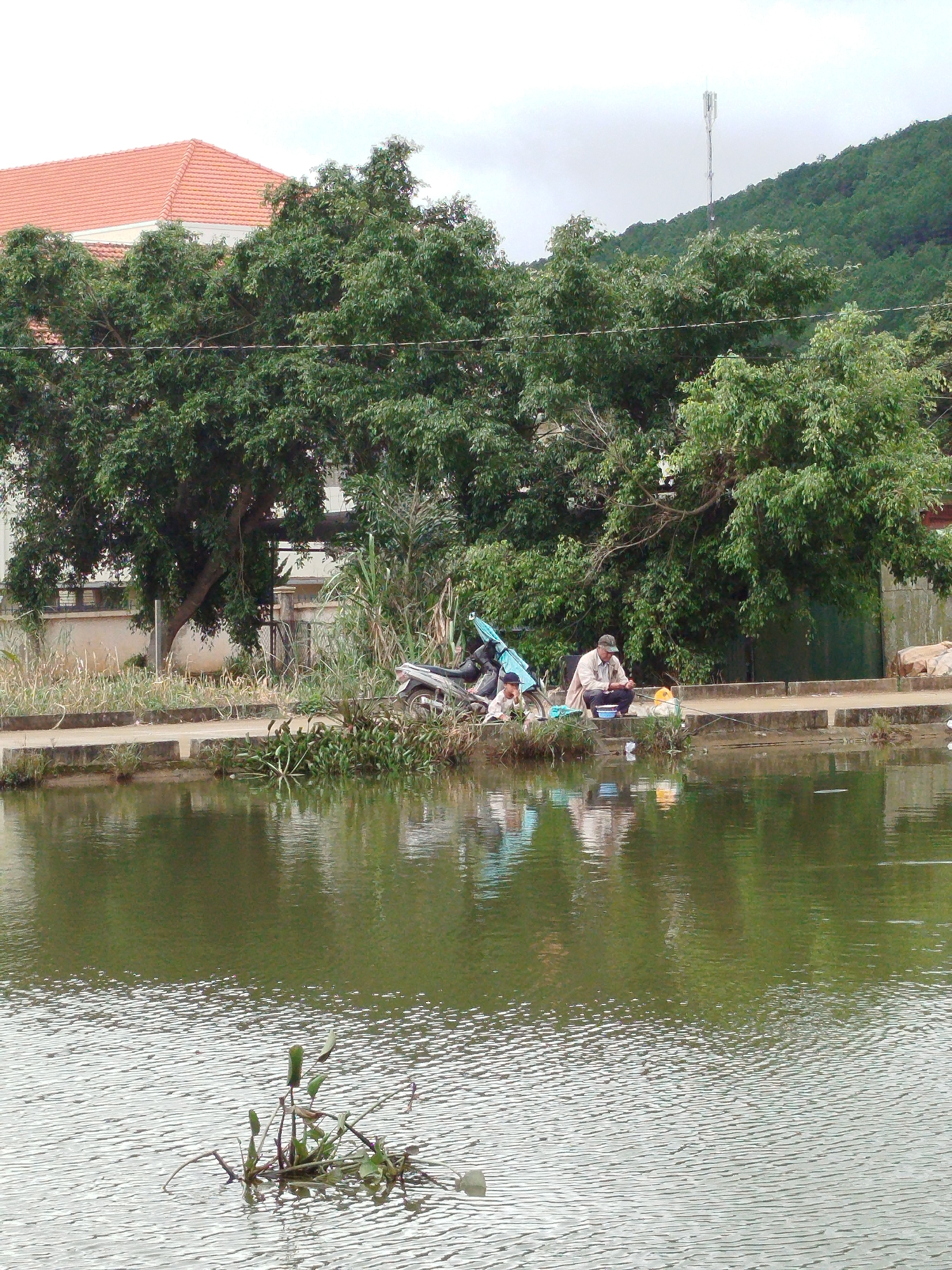 The image captures a waterfront scene in K'Long, Vietnam, focusing on a concrete embankment beside a body of water. In the middle ground, two individuals are seated on the embankment. One person, a male wearing a light-colored jacket and a cap, appears to be fishing with a rod. Beside him is a child, possibly a boy, also seated. A motorcycle is parked behind them, along with some turquoise-colored tarpaulin. The embankment is lined with dry grasses and vegetation, with a larger tree providing shade overhead. In the background, a building with a red tiled roof is visible, partially obscured by dense green foliage. Beyond that, a hilly landscape covered in trees rises, with a telecommunications tower situated on its peak. The water in the foreground is calm, reflecting the surrounding trees and sky, with a small cluster of aquatic plants emerging from the surface. The overall atmosphere suggests a quiet, everyday moment by the water.