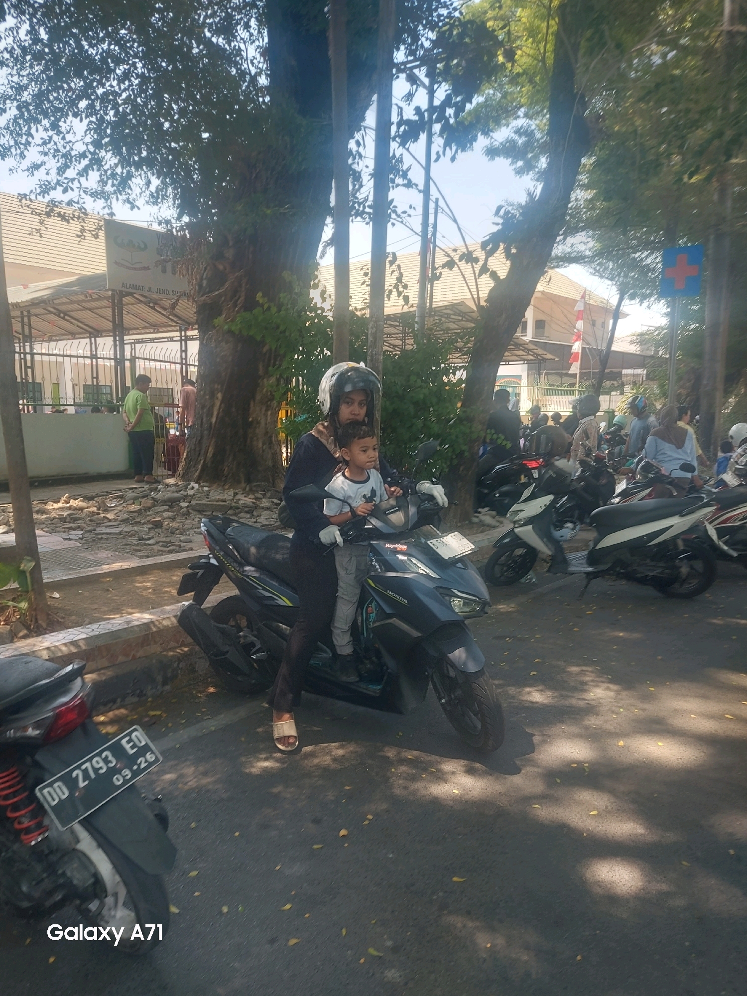 In Makassar, Indonesia, a woman wearing a helmet rides a scooter with a young child seated in front of her. Both are looking forward. The woman is wearing a dark top and dark pants, while the child wears a light-colored t-shirt and gray pants. They are on a street with other motorcycles parked and people in the background. The environment features trees with dappled sunlight on the asphalt and some rubble on the side of the road. A sign with a red cross on a blue background is visible in the distance, suggesting a medical facility or information point. The license plate on the scooter reads "DD 2793 EO".