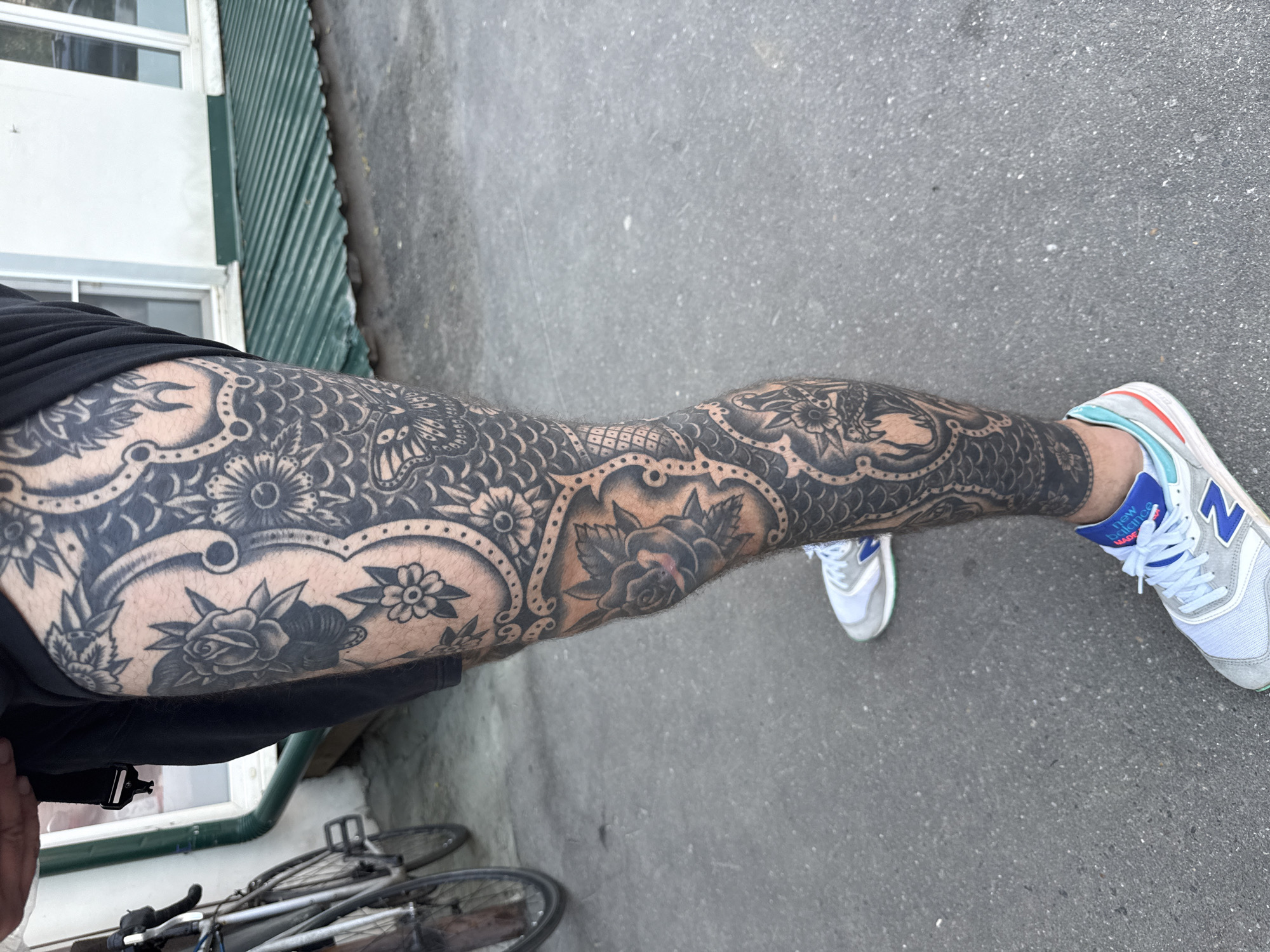 The image shows a person's tattooed leg with a detailed black ink design featuring floral patterns, scales, and possibly mythical creatures, extending from the upper thigh down to the ankle. The person is wearing grey and white New Balance sneakers with blue accents. The leg is positioned on a grey asphalt surface, possibly a sidewalk or street. In the background, there is a bicycle partially visible along with a building with a green corrugated wall and white-framed windows. There is no evidence in the image to suggest the scene is in Moscow, Russia.