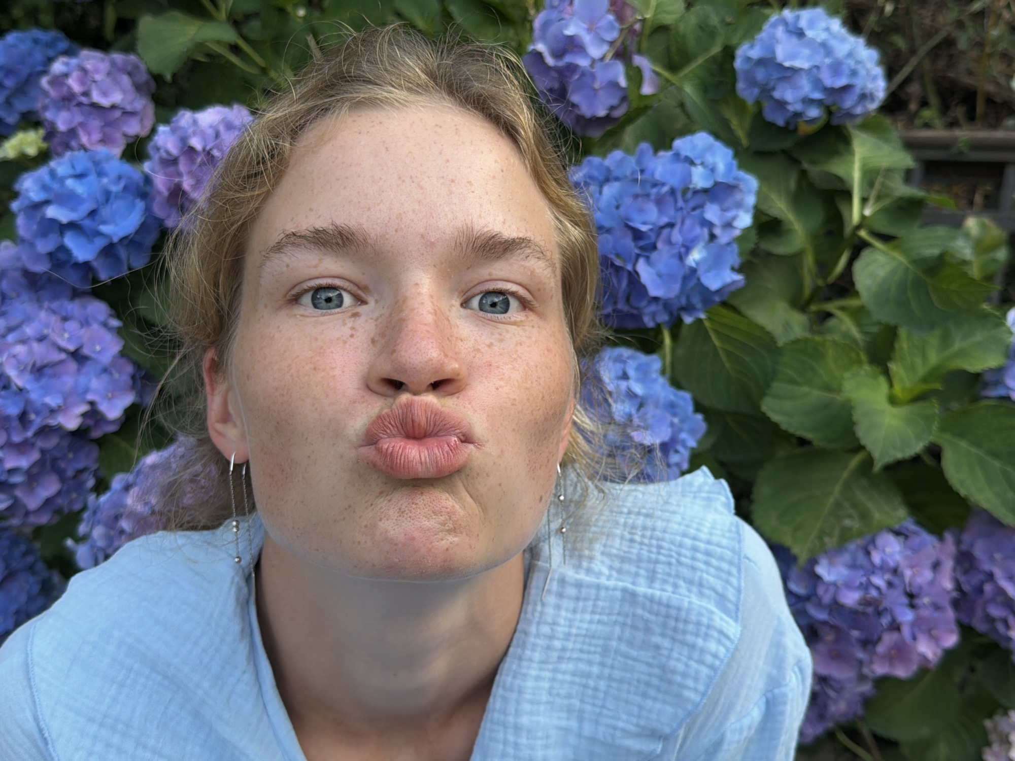 A young woman with blonde hair and blue eyes is in front of a wall of blue and purple hydrangeas. She is wearing a light blue shirt and silver earrings. Her lips are puckered in a kiss. The image appears to have been taken outdoors in Budva, Montenegro.
