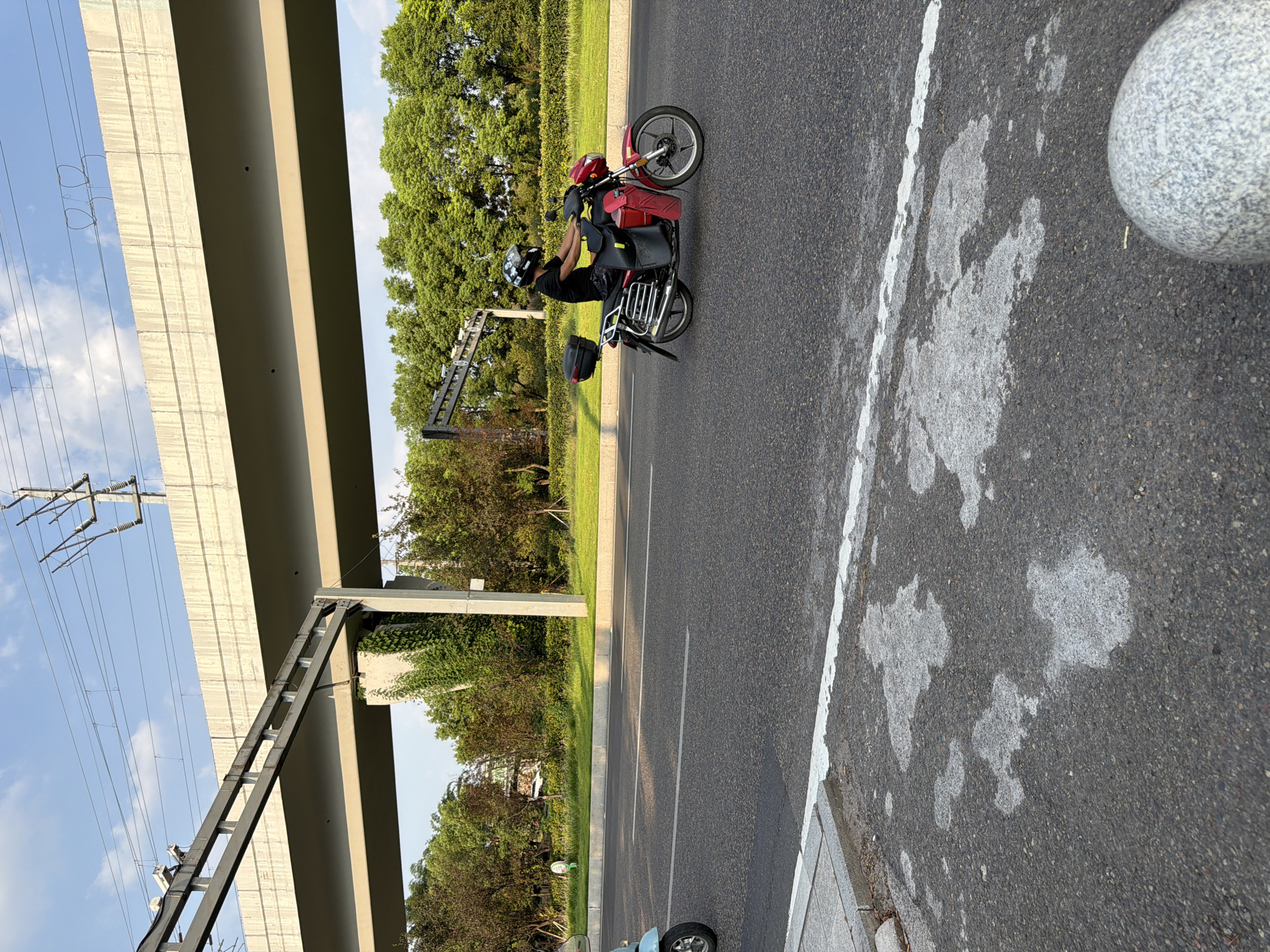 The image shows a person riding a red motorcycle on a road in Jiangjin, China. The motorcycle is a red scooter-style model with a black top case and red saddlebags. The rider is wearing a black t-shirt, dark shorts, and a helmet with a white and black pattern. The road is paved and has white lane markings. In the background, there is an elevated structure, likely a highway overpass, with overhead power lines. Green trees and foliage line the side of the road. The sky is blue with some clouds. The foreground shows a portion of the road surface with some water stains or patches. A small portion of a light blue scooter is visible on the left side of the image. A round, gray, granite bollard is visible in the lower right corner.