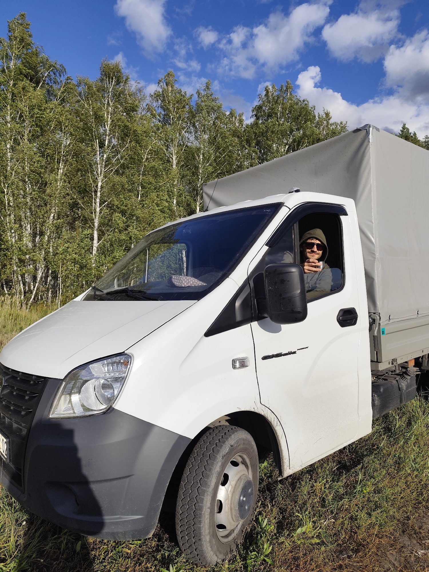 A white, dirty truck is parked on a grassy area next to a forest. A person wearing a green hooded sweatshirt and sunglasses is sitting in the driver's seat and holding a cup. The sky is blue with scattered white clouds. The location is Березов ауылдық округі, Kazakhstan.
