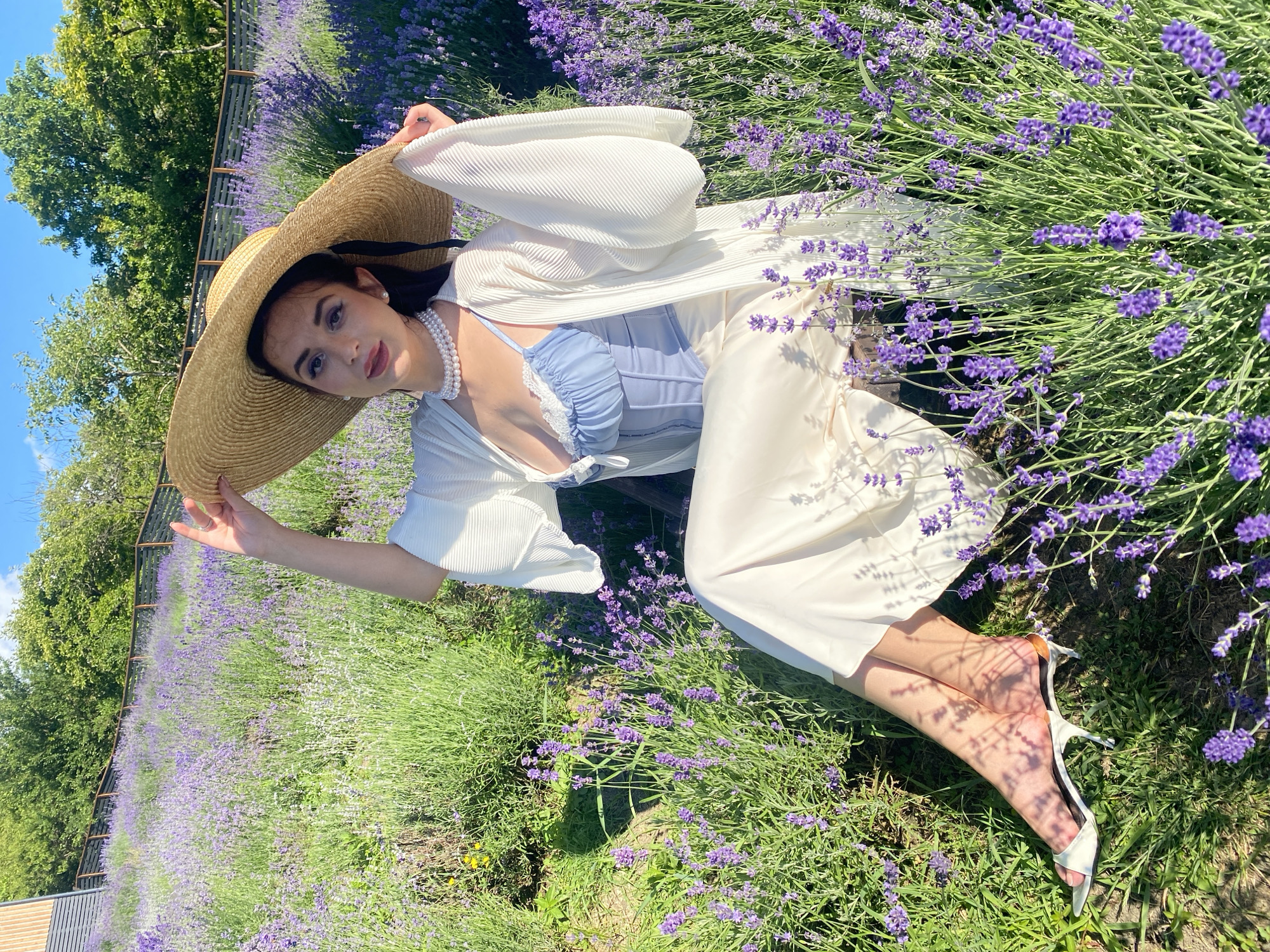 A young woman with dark hair and fair skin lies in a field of lavender, wearing a wide-brimmed straw hat, a white pleated kimono-style robe, a light blue corset top, and white pants. She is adorned with a pearl necklace. Her right hand rests on her hat, and her left hand is not visible. Her legs are crossed at the ankles and she wears white high-heeled sandals. The sun is shining brightly, casting shadows from the lavender plants on her legs. The scene appears to be set in a lavender field, possibly in Verkhneye Uchdere, Russia, on a sunny day. The overall mood is serene and summery. There is no visible text in the image.