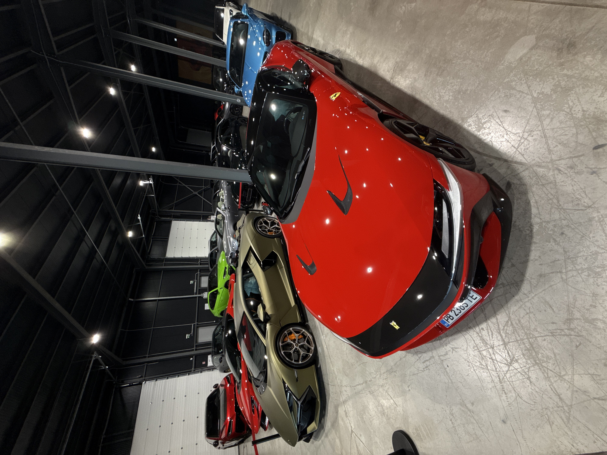 The image shows a collection of luxury sports cars parked indoors, possibly in a showroom or private garage. The cars are predominantly red, gold, and blue. In the foreground, a red Ferrari is prominently displayed. Behind it, a gold Lamborghini is visible. Several other cars are parked in the background, including a bright green Lamborghini, a blue Aston Martin, and various other high-end vehicles. The environment appears to be a large, modern industrial space with a concrete floor and high ceilings. The lighting suggests artificial illumination. The license plate of the red Ferrari is visible and reads "PB 2365 TE". The image quality is sharp and captures the sleek lines of the vehicles. While the image itself does not provide specific information about the location in Plovdiv, Bulgaria, it is presented in a context that suggests it is from there.