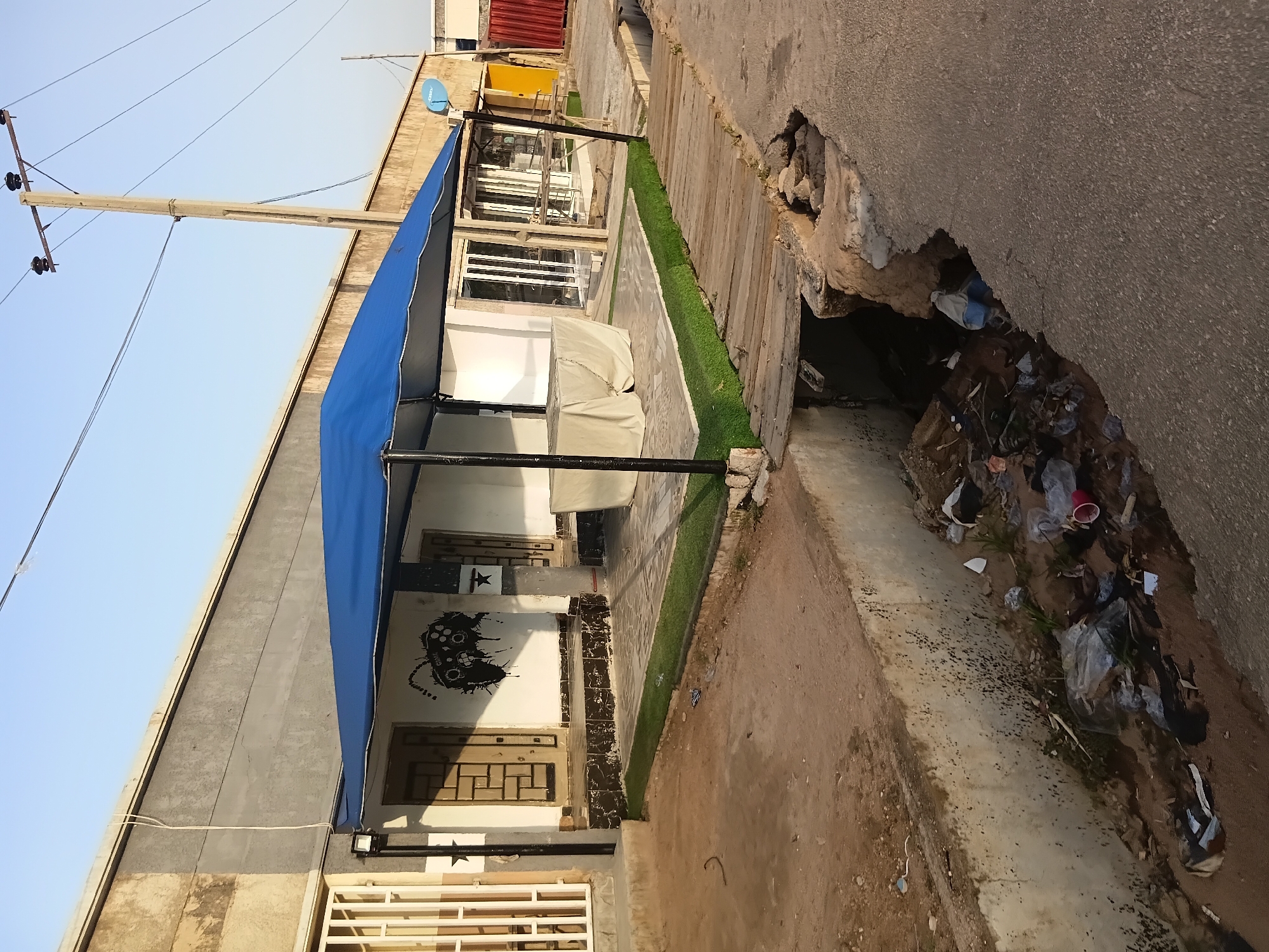 A street-level, exterior, wide shot captures a scene of disrepair and neglect on a sunny day in Jalingo, Nigeria. In the foreground, a dirt path leads to a deep, concrete-lined drainage channel filled with overflowing trash, including plastic bottles and bags. A precarious, makeshift wooden bridge spans the channel, its planks weathered and uneven. Beyond the bridge, a building with a gray facade features a white awning supported by black poles, providing shade over a tiled patio. The patio has a patch of artificial grass and is furnished with a table covered by a white cloth. The building's entrance is marked by patterned doors and a black graphic of a controller on the wall. A power pole with electrical wires stands to the right of the building. In the background, glimpses of other buildings and a blue satellite dish are visible. The overall impression is one of urban decay, with the environment marred by litter and damaged infrastructure.