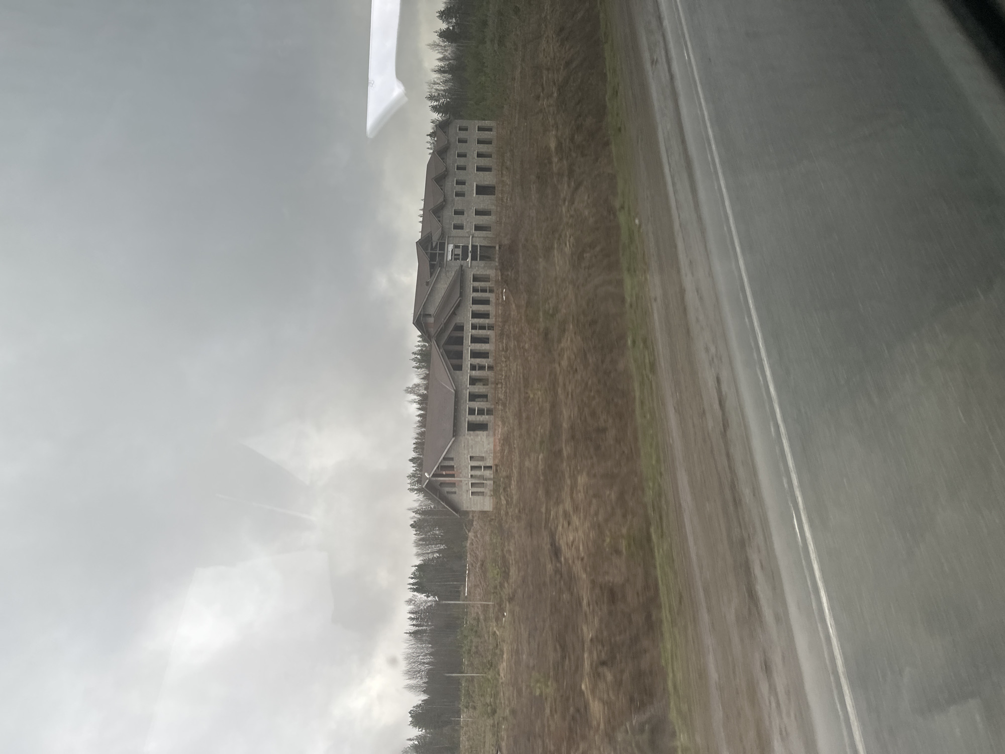 The image depicts an unfinished concrete building, likely a hotel or apartment complex, located in Bolgury, Russia. The structure is multi-storied and shows exposed concrete framing for windows and walls. The surrounding environment appears to be a rural area with a dirt field and a line of trees. An asphalt road runs alongside the building's property. The sky is overcast, suggesting a cloudy or rainy day. The image appears to be taken from inside a vehicle, possibly a bus or car, based on the reflections and the perspective.