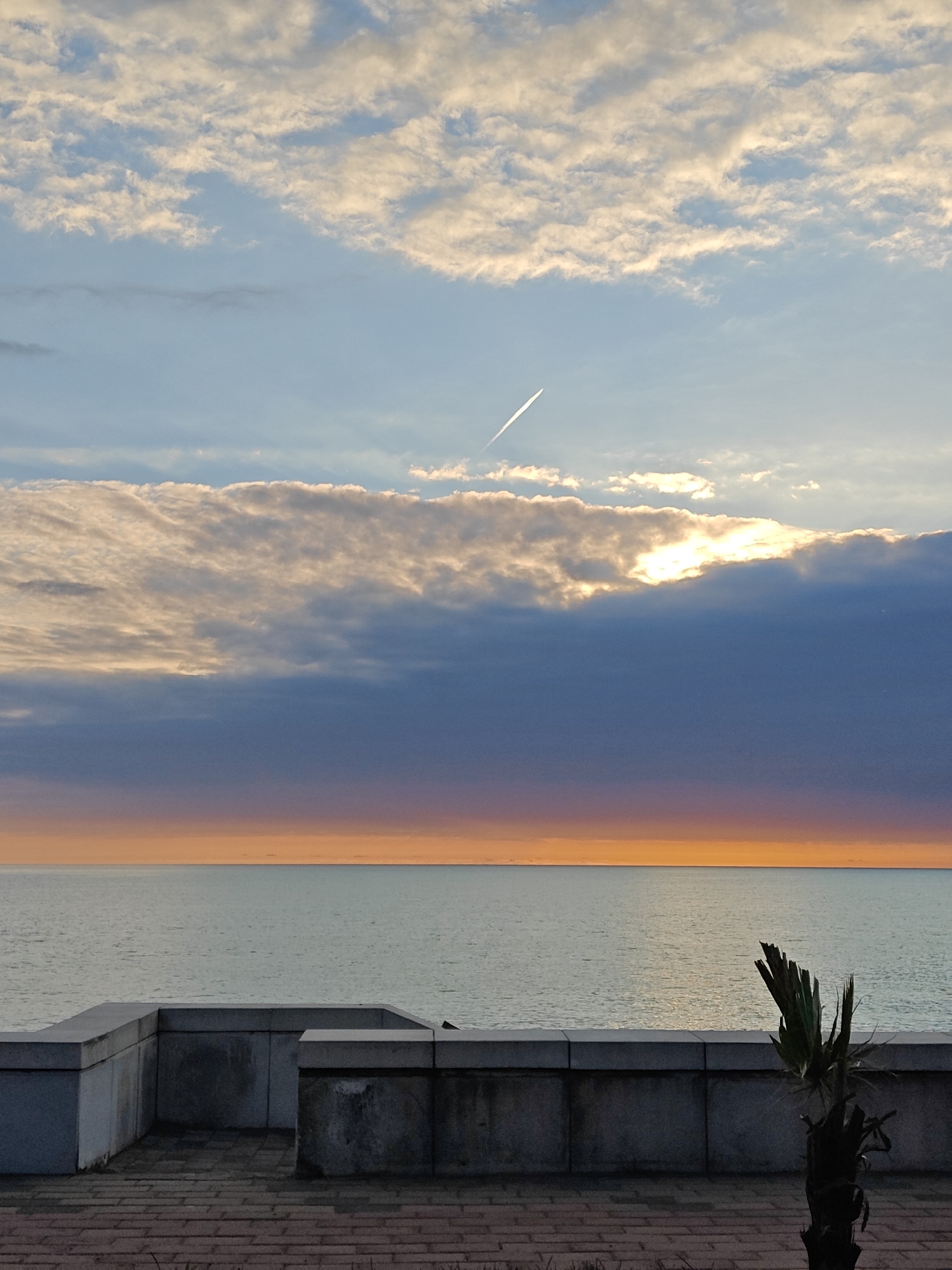 The image displays a serene coastal or lakeside scene captured during either sunrise or sunset. The sky is a prominent feature, showcasing layers of clouds and light. The uppermost part of the sky is a soft blue, adorned with scattered, wispy white clouds. A distinct, thin white airplane contrail diagonally crosses this section from left to right.
Below, a dramatic band of thick, dark clouds stretches across the mid-sky. Through a gap in these heavy clouds on the right, a brilliant burst of sunlight emerges, creating a striking contrast. Just above the horizon, a vibrant orange-yellow glow casts a warm hue over the expansive, calm body of water, suggesting the sun's position. The water itself reflects the muted colors of the sky and horizon.
In the foreground, a grey, angular concrete structure, possibly a barrier or part of a promenade, runs horizontally, overlooking the water. The ground beneath it is paved with reddish-brown interlocking bricks or tiles. On the far right, a small, dark silhouette of a palm-like plant adds a touch of vegetation to the scene. No human activity or visible text is present, and the overall atmosphere is one of peaceful natural beauty.