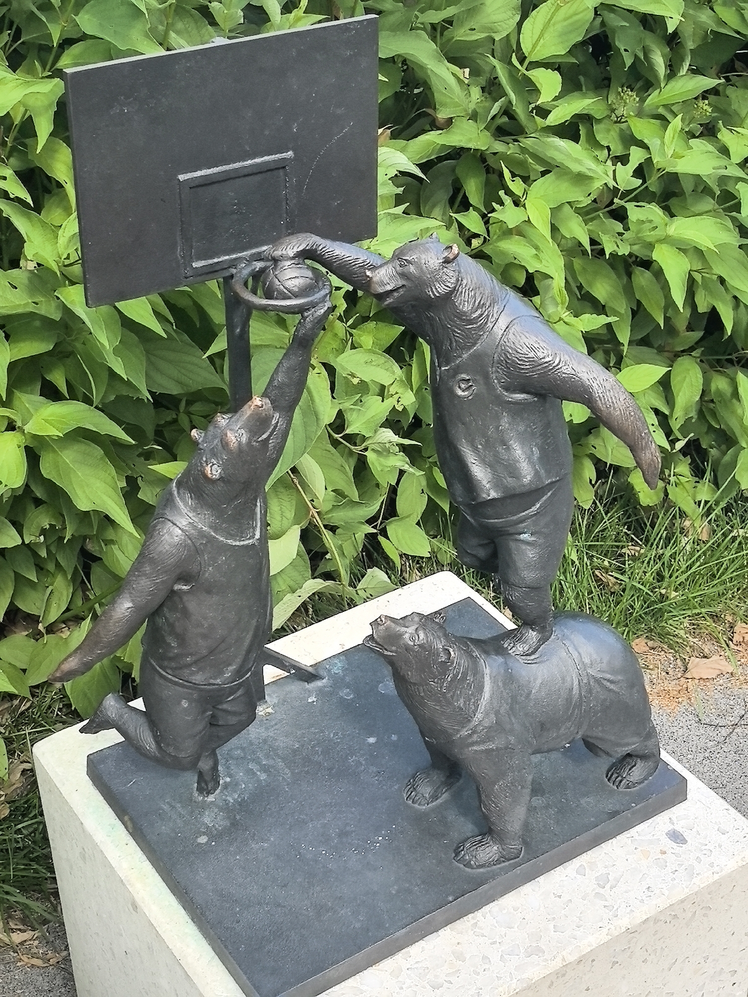 A bronze sculpture depicts three bears playing basketball. Two bears are standing, one on top of the other, reaching for a basketball near a backboard. The bottom bear stands on its hind legs, supporting the bear above it. The bear on top is reaching for the basketball with one paw, while its other paw is positioned behind the ball. A third bear is positioned lower, on the ground, looking up at the action. The sculpture is situated outdoors, with lush green foliage in the background. This sculpture is located in Novomoskovsk, Russia.