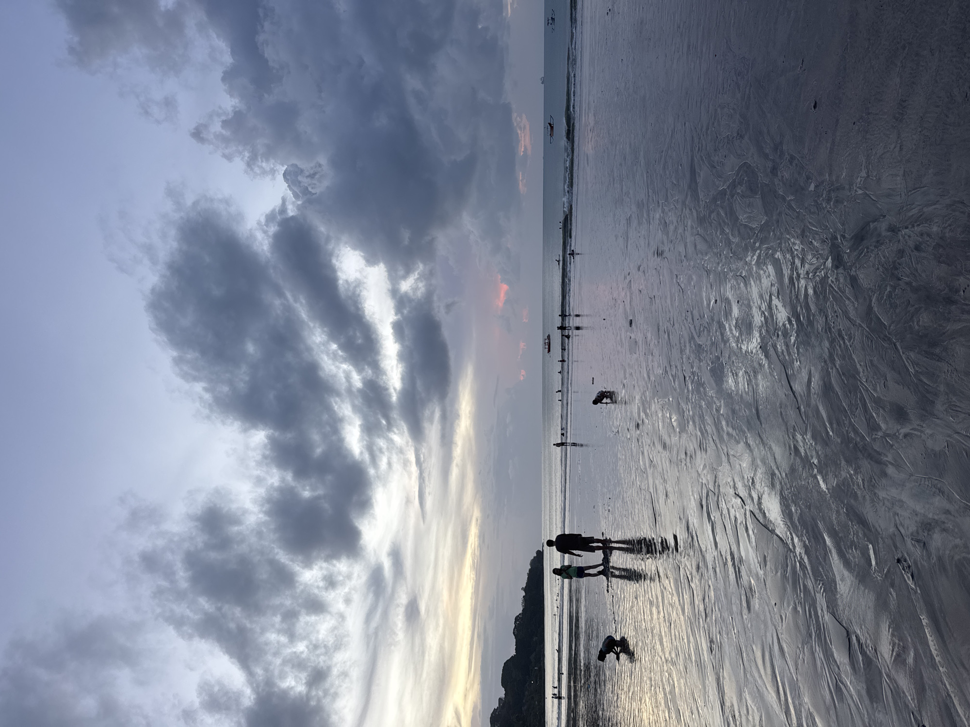 The image captures a serene coastal scene at either sunrise or sunset, with a dramatic sky dominating the upper portion. The sky is largely overcast with large, dark grey clouds, particularly dense towards the upper left. However, streaks of lighter, more elongated clouds nearer the horizon reveal soft hues of pale orange and pink, hinting at the sun's low position.
Below the horizon, a vast expanse of wet, reflective sand stretches into the foreground, showing intricate patterns and ripples left by the receding tide. The wet sand brilliantly mirrors the sky's light, creating elongated silhouettes of people and animals.
Several figures are scattered across the beach. In the foreground, two silhouetted individuals, possibly a couple, walk close together, their long shadows stretching out behind them. Further back, another person is seen bending down, possibly interacting with a small dog beside them. Another dog is visible alone on the sand further in the distance. More distant figures, some possibly with surfboards, are visible near the water's edge, suggesting activities like surfing or wading. Small boats are also dotted on the calm ocean surface near the horizon.
The overall atmosphere is peaceful and contemplative, indicative of leisure time spent enjoying the natural beauty of the beach during the golden hour. No visible text is present in the image.