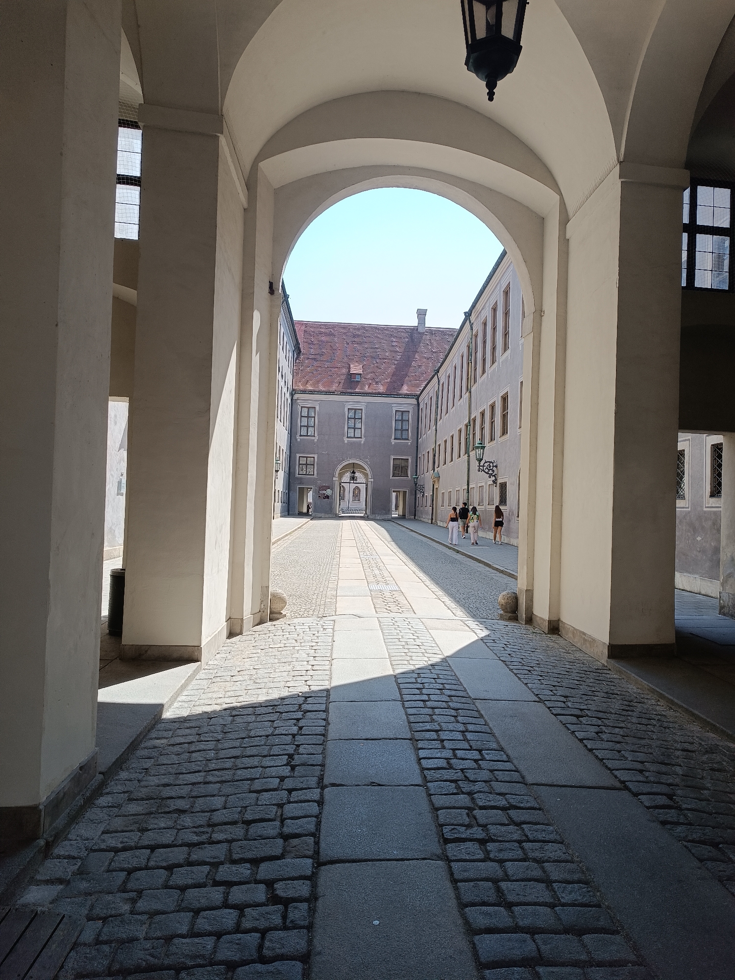 The image depicts a sun-drenched cobblestone pathway leading through a covered archway in Munich, Germany. The architecture suggests a historical setting, possibly part of a palace or significant building complex. The pathway is lined with light-colored stone slabs and darker cobblestones, creating a geometric pattern. Sunlight casts strong shadows across the ground, indicating a bright, clear day.
On the right side of the pathway, a long, multi-story building with numerous windows and a red-tiled roof is visible. Several people are walking away from the viewer on this side of the pathway, casually dressed. Two figures in the foreground on the right are obscured by the shadows cast by the archway columns. On the left side of the pathway, another building is partially visible, also with windows and a red roof.
An ornate black lantern hangs from the curved ceiling of the archway, illuminated by the ambient light. The archway itself is constructed of pale, smooth stone, with large, rounded arches forming the primary structure. The overall scene conveys a sense of historical grandeur and a typical summer day in a European city. The precise location is identifiable as the Marstall courtyard of the Munich Residenz.