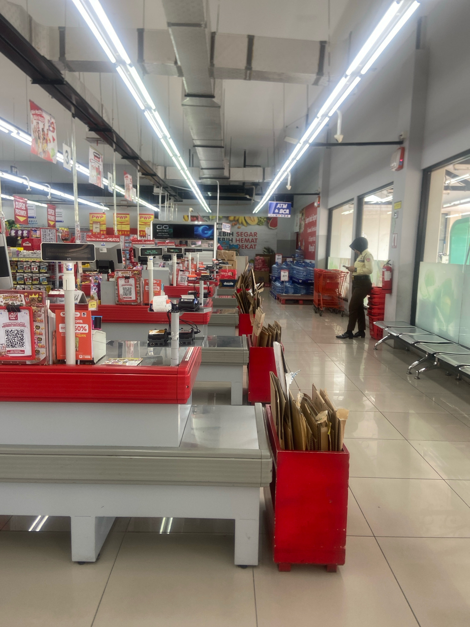 The image shows the interior of a supermarket in Ngaglik, Indonesia. The perspective is from behind a row of checkout counters, looking down an aisle of the store.
The foreground is dominated by the red and white structure of the checkout counters. Several of these counters are visible, each with a white base and a red top surface. On the red surfaces, various items are placed, including small black receipt printers, white screens, and various signs. One counter has a QR code displayed on a white sign with red lettering. To the right of the counters, there are red bins filled with brown paper bags.
In the middle ground, more checkout counters are visible, as well as shelves stocked with products. Signs with red and yellow lettering hang above some of the shelves. Further down the aisle, there are stacks of bottled water on pallets. A red shopping cart is also visible.
The background shows the ceiling with fluorescent lighting and exposed ductwork. To the right, there is a glass partition, behind which a security guard in a light brown uniform and dark headscarf is standing, looking at their phone. Next to the guard is a red fire extinguisher. Further to the right, there are several metal benches. The floor is tiled with a beige color. The overall environment appears to be a well-lit and organized supermarket.