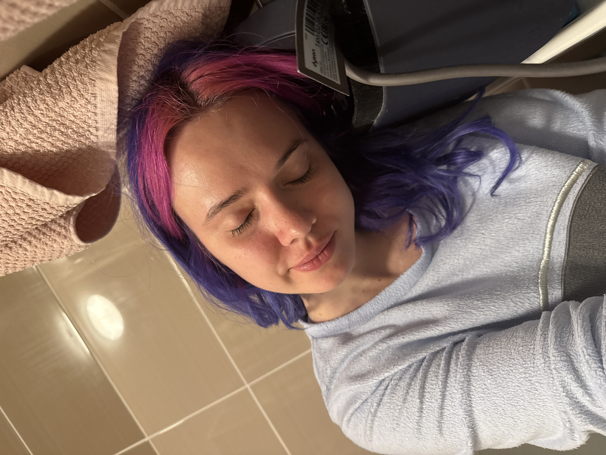 A medium shot from an overhead perspective captures a young woman lying down indoors, appearing to be resting or sleeping with her eyes closed. She has distinctive bi-colored hair, transitioning from vibrant pink at the roots to purple along the lengths. Her facial features are relaxed, and she has fair skin with a slight sheen on her forehead.
She is dressed in a light blue, fuzzy long-sleeved top or sweater. To her left, a light beige or pinkish terry cloth towel is bunched up near her head. Behind her head, a dark object is partially visible, featuring a white label with the text "Dyson" and "TAN" clearly legible. A thick, dark gray cable extends from this object.
The background consists of light-colored, possibly beige or cream, square tiles with visible grout lines. A prominent, bright, oval-shaped reflection, likely from an overhead light fixture, is seen on the tiled surface. The scene suggests a private, indoor setting, possibly a bathroom or a similar tiled room. The woman's posture and closed eyes convey a sense of peaceful relaxation. The image itself does not contain specific visual cues to indicate the location is Zagreb, Croatia.