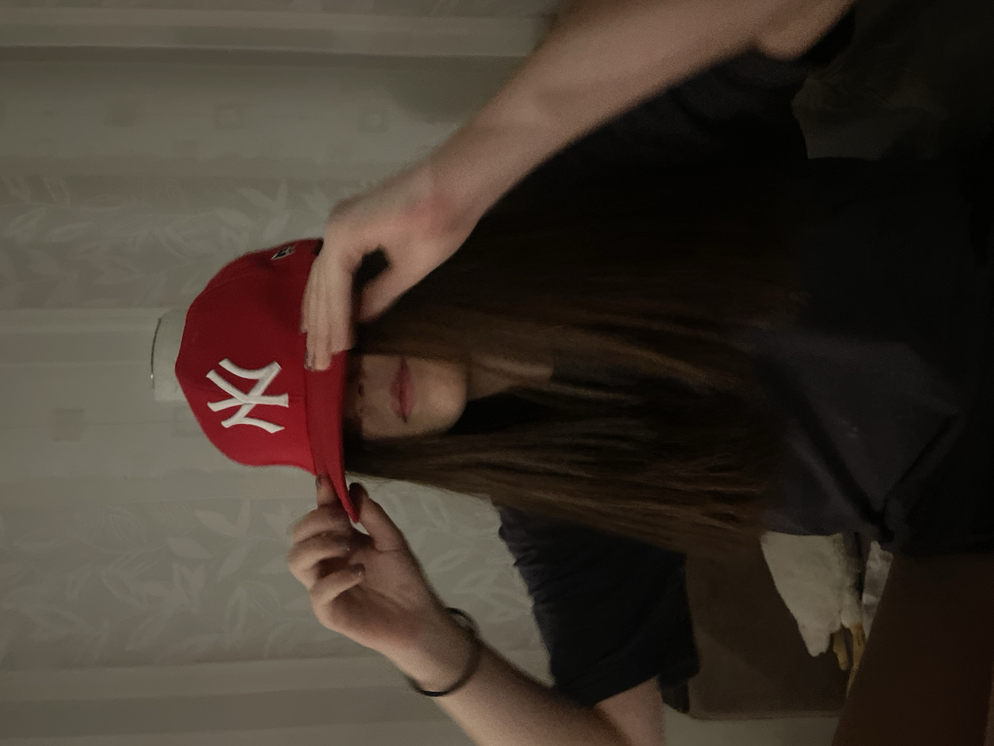 A person with long brown hair is wearing a red New York Yankees baseball cap. The person is wearing a black t-shirt. The background is a white wall with a patterned wallpaper. The location is potentially Murino, Russia, but this cannot be definitively determined from the image.