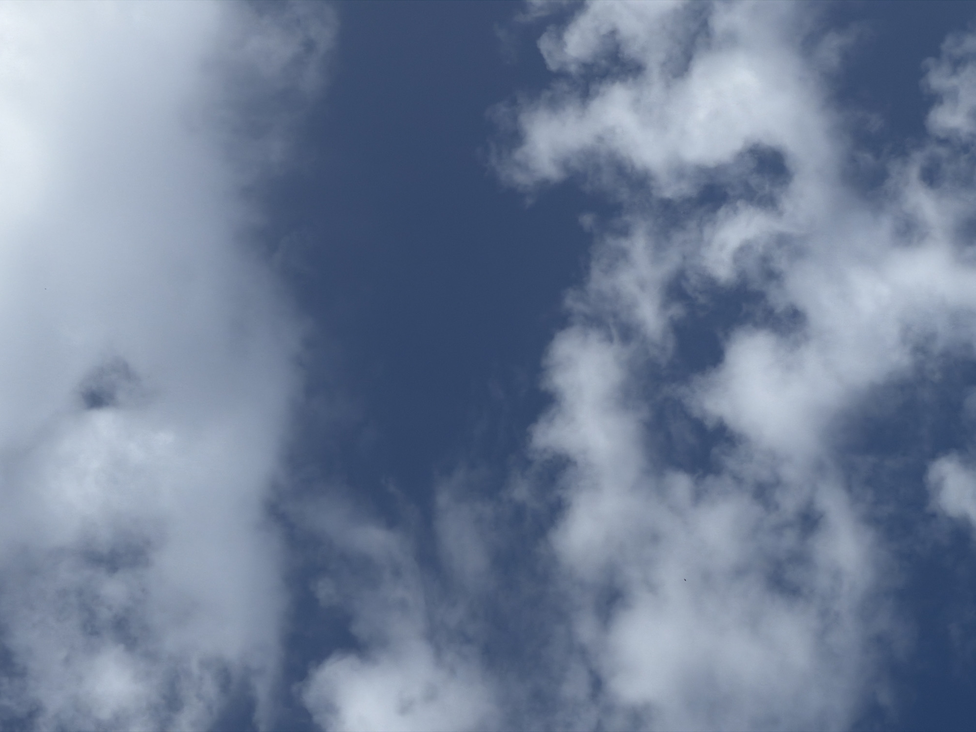 The image depicts a close-up view of a cloudy sky. The clouds are predominantly white and grey, with varying densities, suggesting a mix of cumulus and stratus formations. The sky itself is a deep blue, indicating daylight conditions. The composition is abstract, focusing on the textures and patterns of the clouds against the blue expanse. There are no discernible city landmarks or specific locations within Barcelona visible in this image. The scene is entirely composed of atmospheric elements.