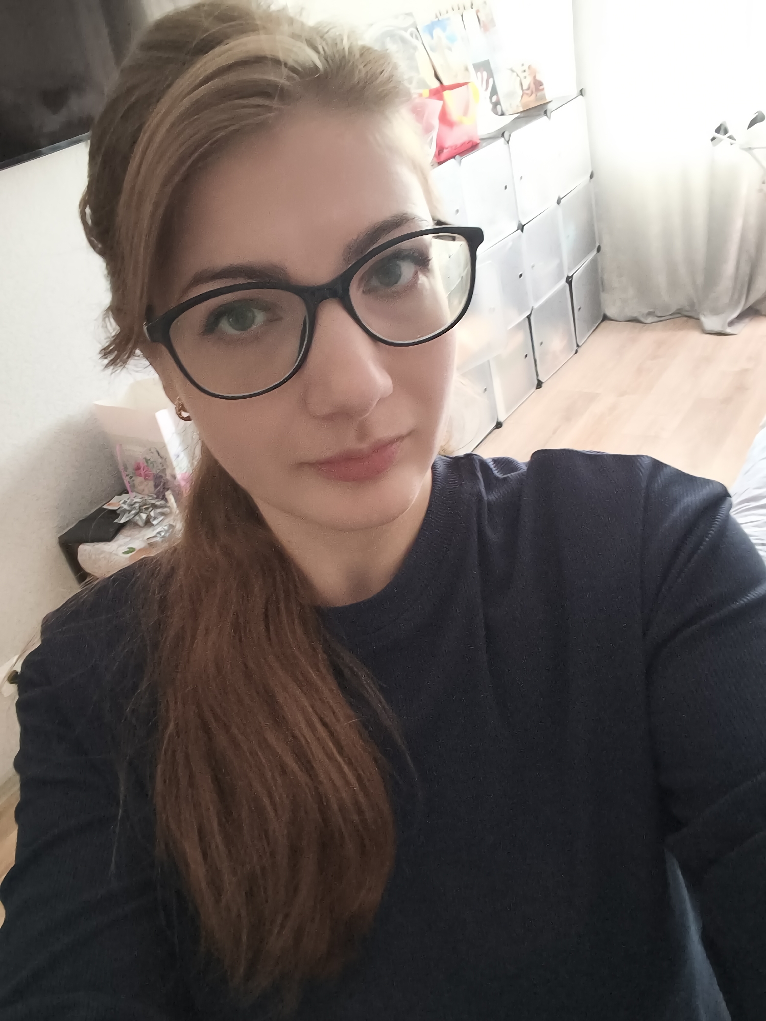 A young woman with blonde hair pulled into a ponytail and wearing black-rimmed glasses is looking at the camera. She is wearing a dark blue sweater and has a slight smile on her face. She is in an indoor setting with a white wall and light-colored flooring. To her left, there are shelves filled with various items, including what appear to be books or boxes. To her right, there is a white curtain. The overall lighting suggests the photo was taken during the day.