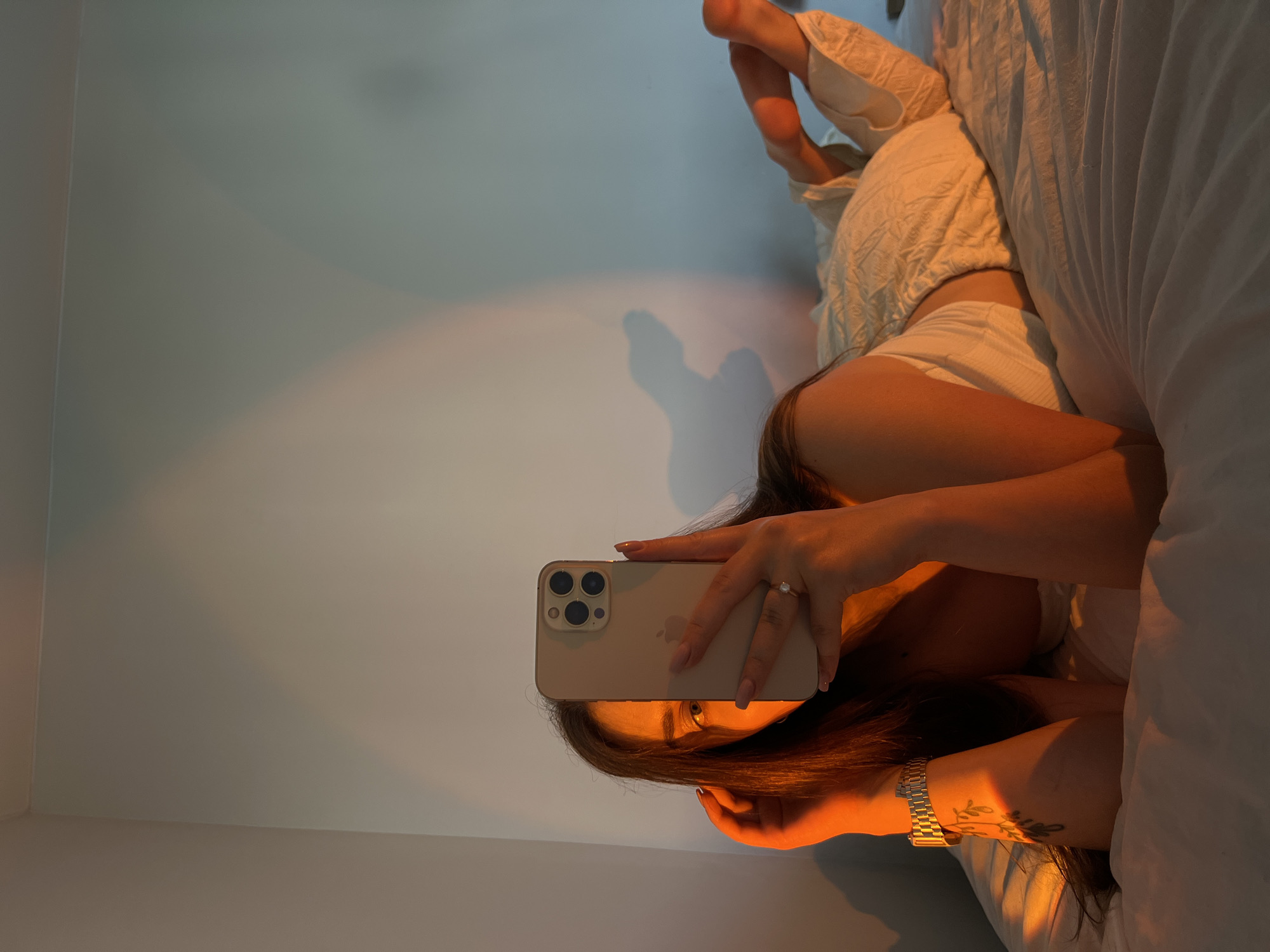 A young woman is lying on a bed, taking a selfie with her phone. She is partially covered by the phone, but her eye is visible. The room is lit by warm, golden light, casting shadows on the wall. The woman is wearing a white tank top and light-colored pants. She has a watch on her left wrist and a ring on her right hand. The bed appears to be in a bedroom. The image does not contain any specific details that would allow for identification of the city of Ternopil, Ukraine, or any other location.