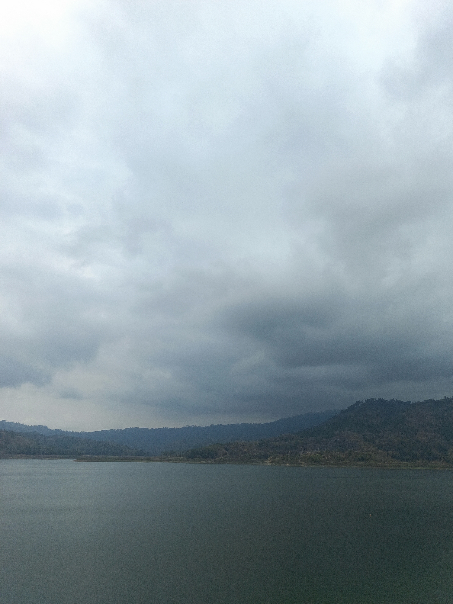 The image depicts a wide, still body of water stretching across the foreground, reflecting the overcast sky. In the mid-ground, a low, tree-covered shoreline is visible, leading into rolling hills and mountains in the background. The landscape is predominantly muted in color, with shades of gray, blue, and dark green dominating the scene. The sky is filled with heavy, dark clouds, suggesting an impending storm or generally gloomy weather. The overall impression is one of natural tranquility, with no human presence or man-made structures readily apparent. The location, identified as Segawe, Indonesia, suggests a rural or natural setting within that region.