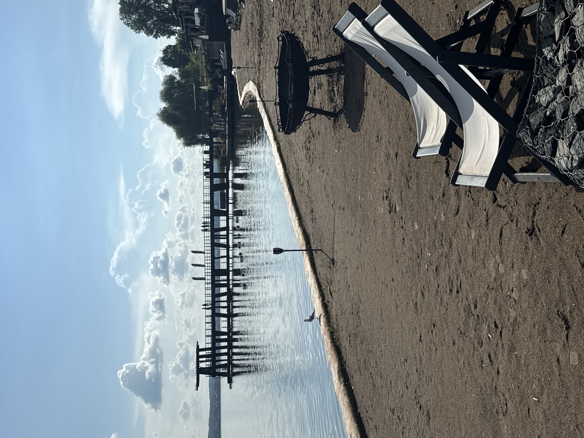 The image depicts a waterfront scene with a sandy beach in the foreground. Two white deck chairs are positioned on the sand, facing away from the viewer. To the right of the chairs, a large, black metal fire pit with a wide basin and three legs stands on the sand. A curved, sandbag barrier separates the beach from the water. A seagull stands on this barrier near the water's edge. In the middle ground, a long wooden pier extends into the calm, rippling water. Several people are visible on the pier, walking and standing. The pier has a dark, silhouetted structure at its far end. The background features lush green trees and some buildings on the far bank of the water. The sky is a vibrant blue with scattered white cumulus clouds. The overall impression is a serene, sunny day at a recreational waterfront area, likely in Votkinsk, Russia, given the context of the prompt.