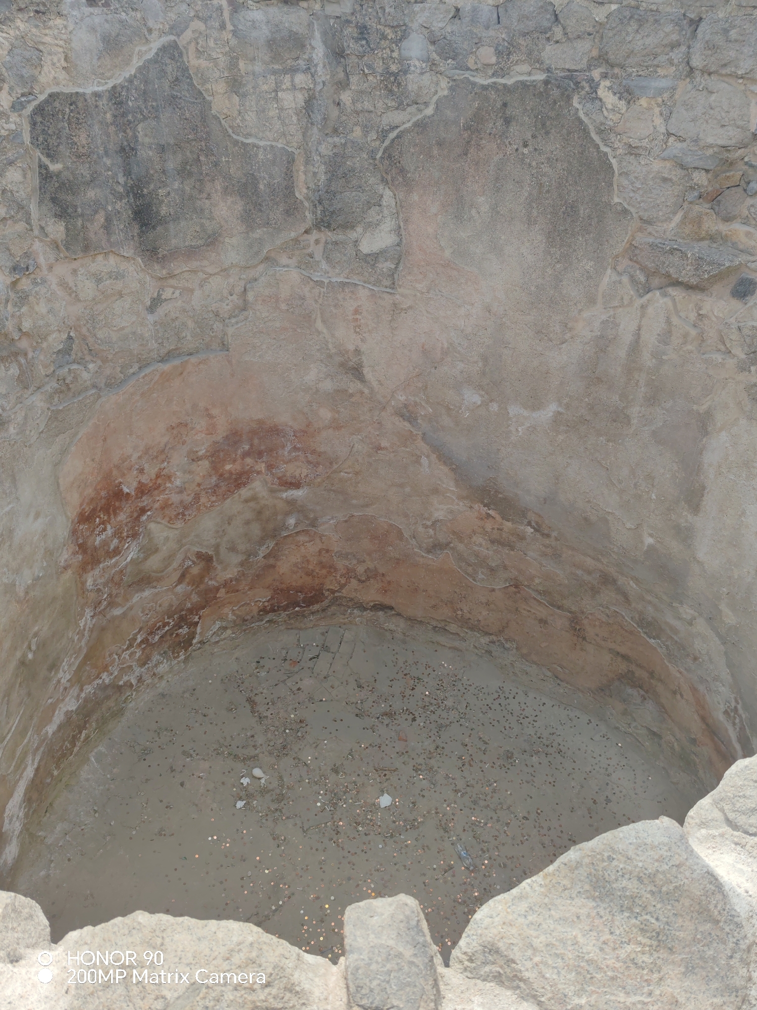 The image depicts the interior of a partially exposed ancient stone well, located in Plovdiv, Bulgaria. The walls of the well are constructed from rough-hewn stone blocks. The interior surface shows signs of wear and staining, with a notable horizontal band of reddish-brown discoloration around the well's circumference. The bottom of the well is visible and contains a scattering of coins. The image is taken from an elevated vantage point looking directly into the well. A portion of the outer stone structure is visible in the foreground. The image includes a watermark in the lower left corner "HONOR 90 200MP Matrix Camera".