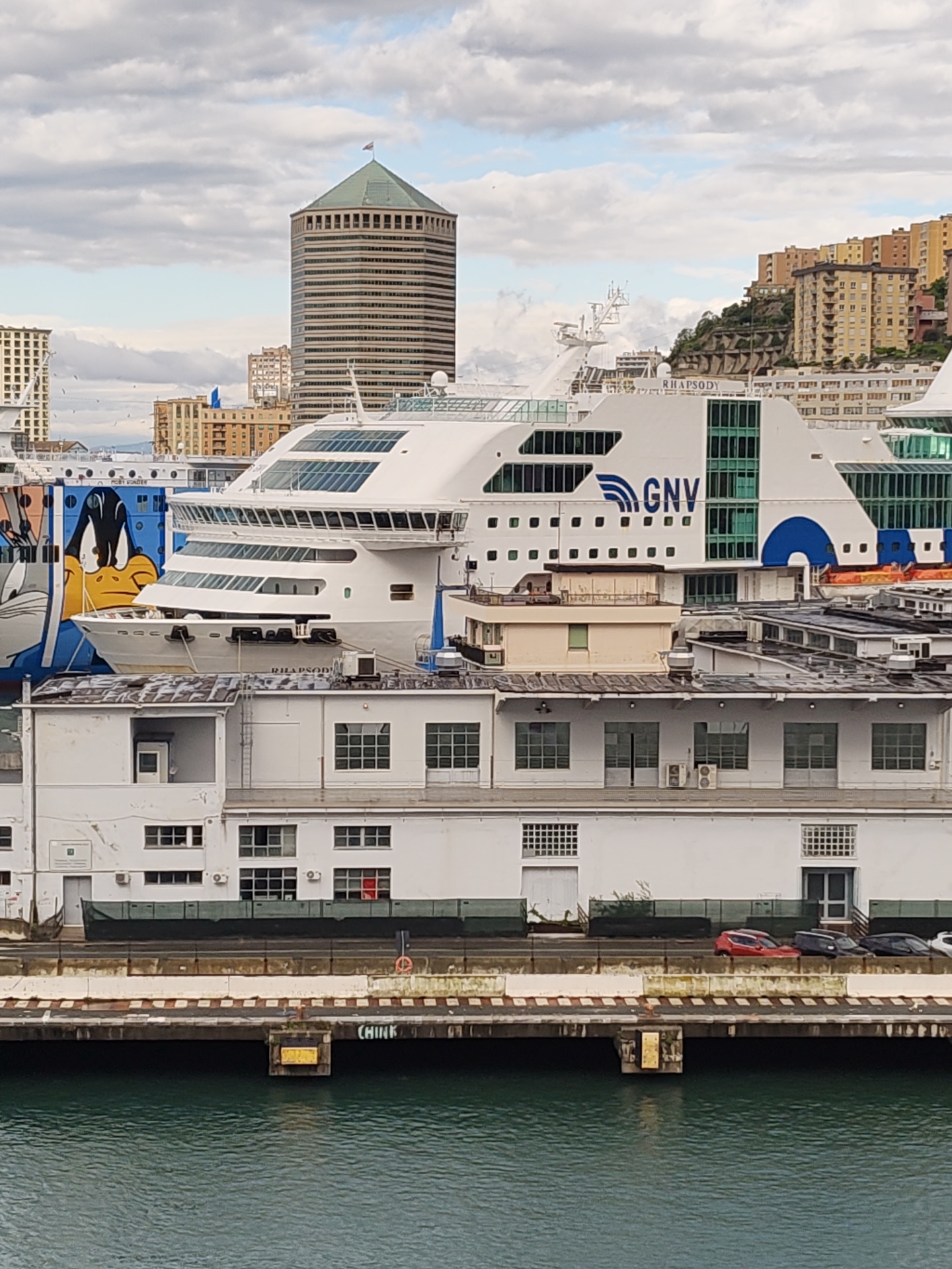 The image depicts a harbor scene in Genoa, Italy. In the background stands the Torre Matitone, a distinctive cylindrical skyscraper with a green roof. A large white ferry ship, with "GNV" emblazoned on its side, is docked alongside a pier. Another vessel with a cartoon graphic is partially visible to the left. The pier features a white building with multiple windows, and several cars are parked along the waterfront. The water appears calm. The sky is overcast with scattered clouds.