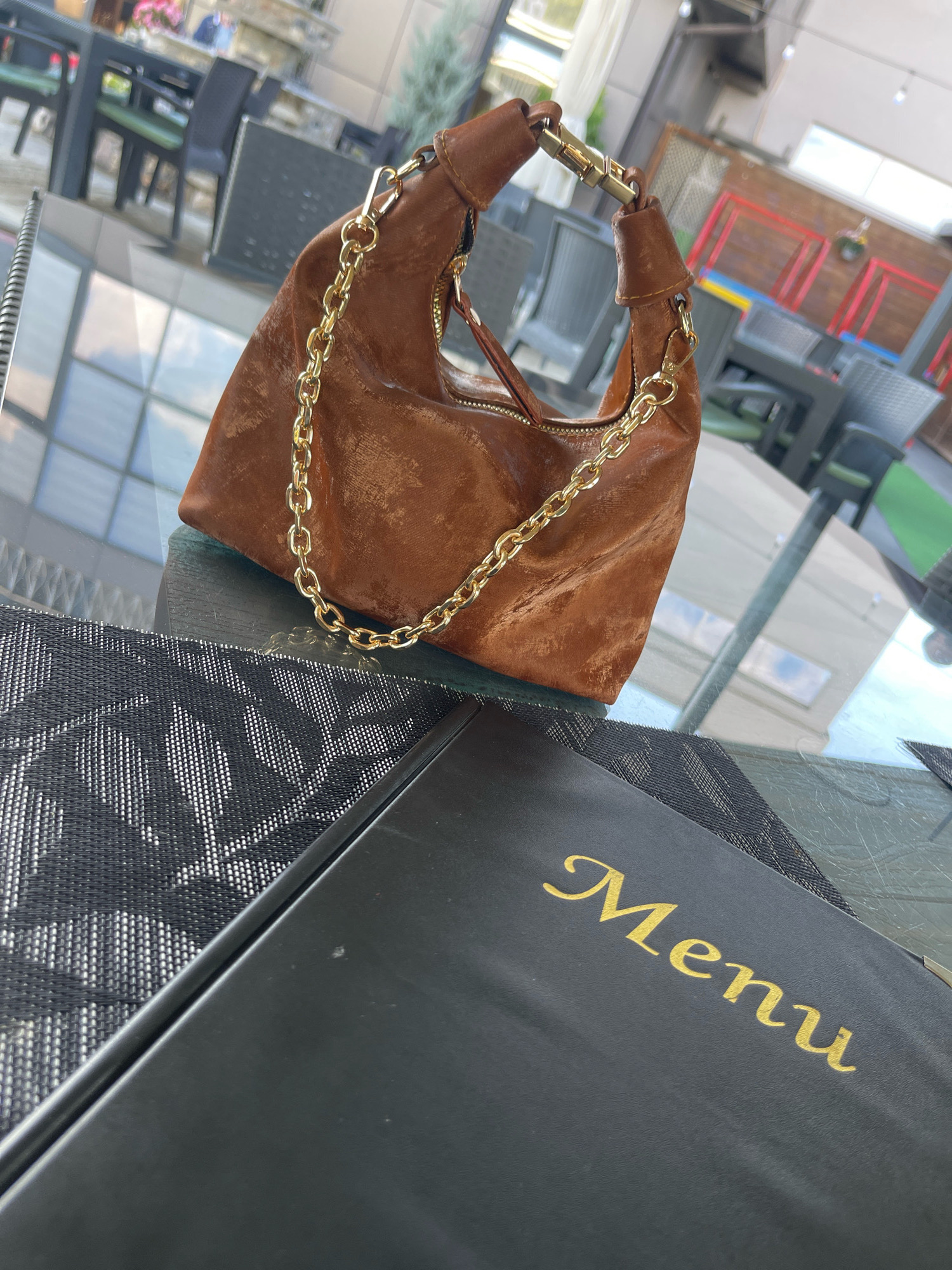 A brown leather purse with a gold chain strap rests on a glass table next to a black menu with gold lettering that reads "Menu." The table is set on a terrace of an outdoor restaurant in Mirushë, Kosovo. In the background, several empty tables and chairs are visible, along with a glimpse of a red railing and some greenery. The lighting suggests it is daytime.