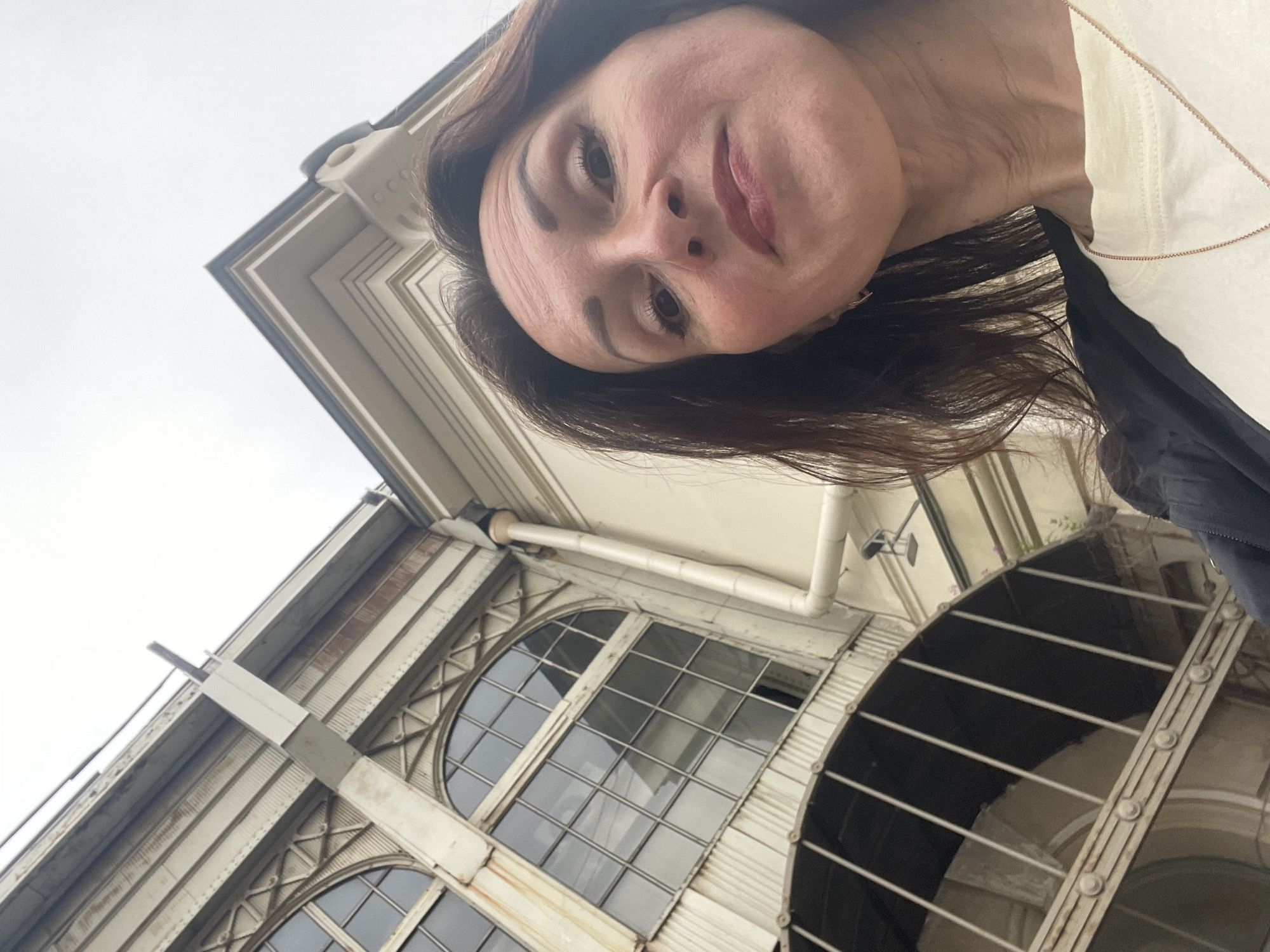 A woman takes a low-angle selfie in front of an old industrial building in Saint Petersburg, Russia. The building has large arched windows with a grid of panes and metal supports. A white downspout runs vertically down the side of the building. The woman has long, dark brown hair and is wearing a light-colored t-shirt and a dark jacket. The sky is overcast and the overall lighting is dim.