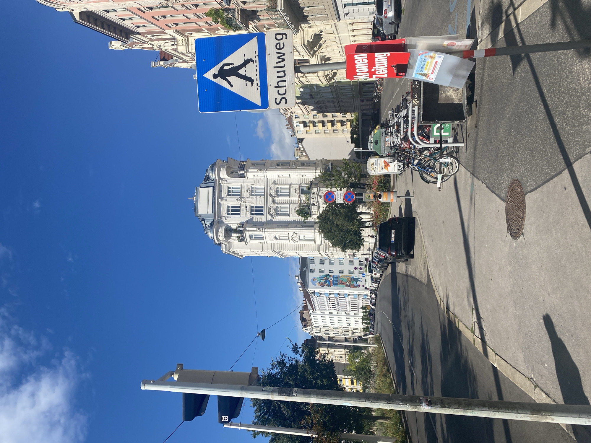 The image captures a street scene in Vienna, Austria, on a bright, sunny day with a clear blue sky. The perspective is from the sidewalk looking down a moderately wide street lined with buildings and parked cars.
In the foreground, a gray concrete sidewalk leads towards the street. A lamppost stands on the sidewalk, and to its right is a blue traffic sign indicating a pedestrian crossing ("Schulweg" in German, meaning school route). Below this sign, a red kiosk displays a newspaper. Further to the right, a row of parked bicycles is visible.
The street itself is paved and curves gently to the left. It is lined with parked cars on the right side. The buildings flanking the street are characteristic of Viennese architecture, with ornate facades, some painted in light colors like white and light pink, and others in muted tones. The building in the center of the background is a prominent white structure with a rounded, domed top.
In the distance, the street continues, with more buildings and trees visible. The overall impression is a well-maintained urban environment. There are no people clearly visible in the image, and no specific actions are taking place.