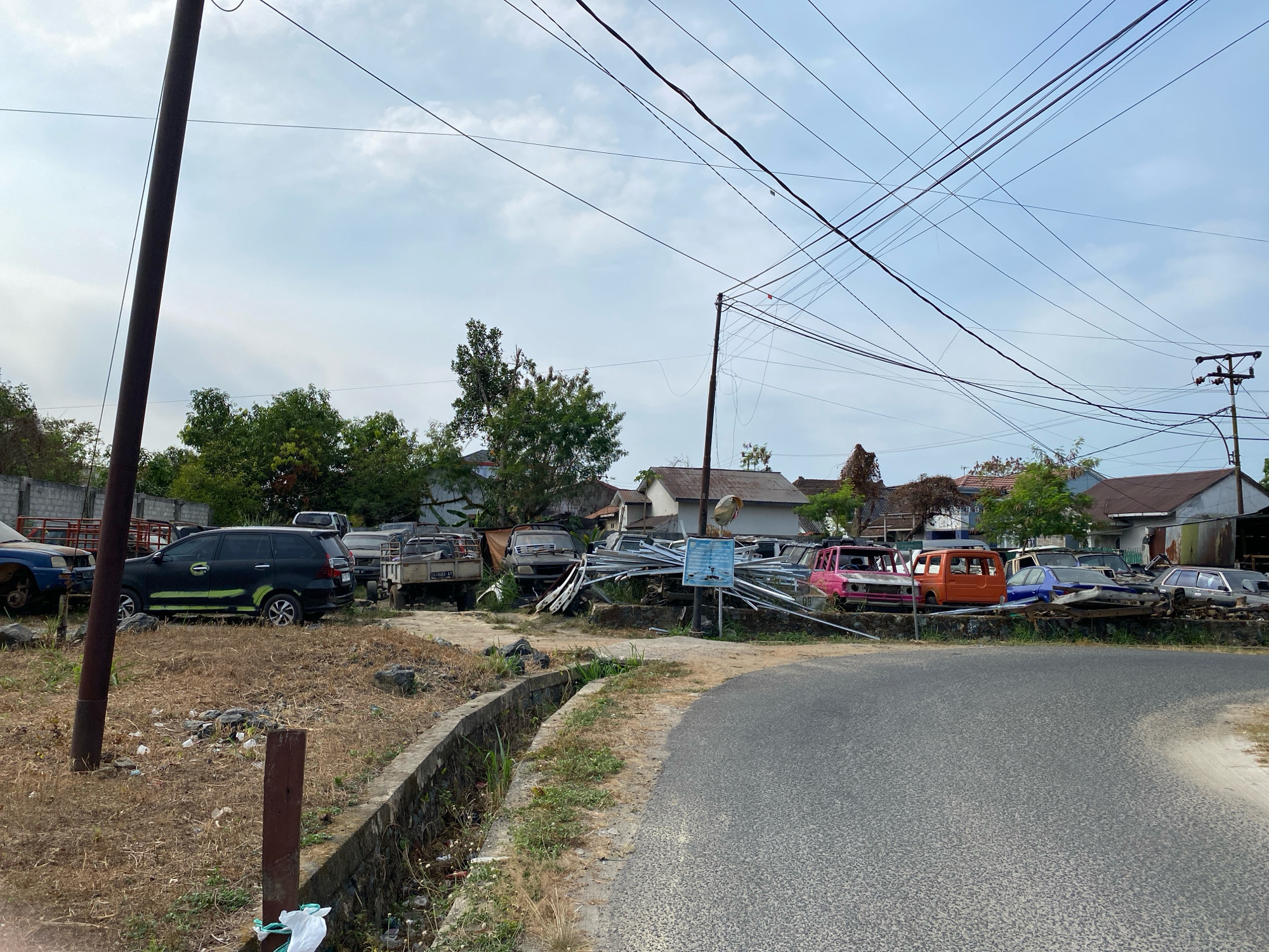 The image depicts a rural roadside scene in Banjarbaru, Indonesia, characterized by an accumulation of abandoned vehicles and construction materials. In the foreground, a paved road curves to the right, bordered by a concrete drainage ditch. Dry, overgrown grass lines the ditch and the unpaved area adjacent to the road. Several wooden posts stand in the unpaved area, one with a plastic bag caught on it.
The primary focus of the image is a collection of dilapidated vehicles parked along the roadside. These include a black minivan with green markings on its sides, a small truck with a license plate starting with "LG 4803 BF", a grey pickup truck, and several other cars in various states of disrepair, some appearing to be stripped of parts. Among the vehicles, there are also stacks of metal pipes and other construction materials. In the background, houses with corrugated metal roofs and trees are visible under a cloudy sky. Overhead, a dense network of electrical wires crisscrosses the sky, supported by utility poles. The overall impression is one of disuse and abandonment. There are no people visible in the image.