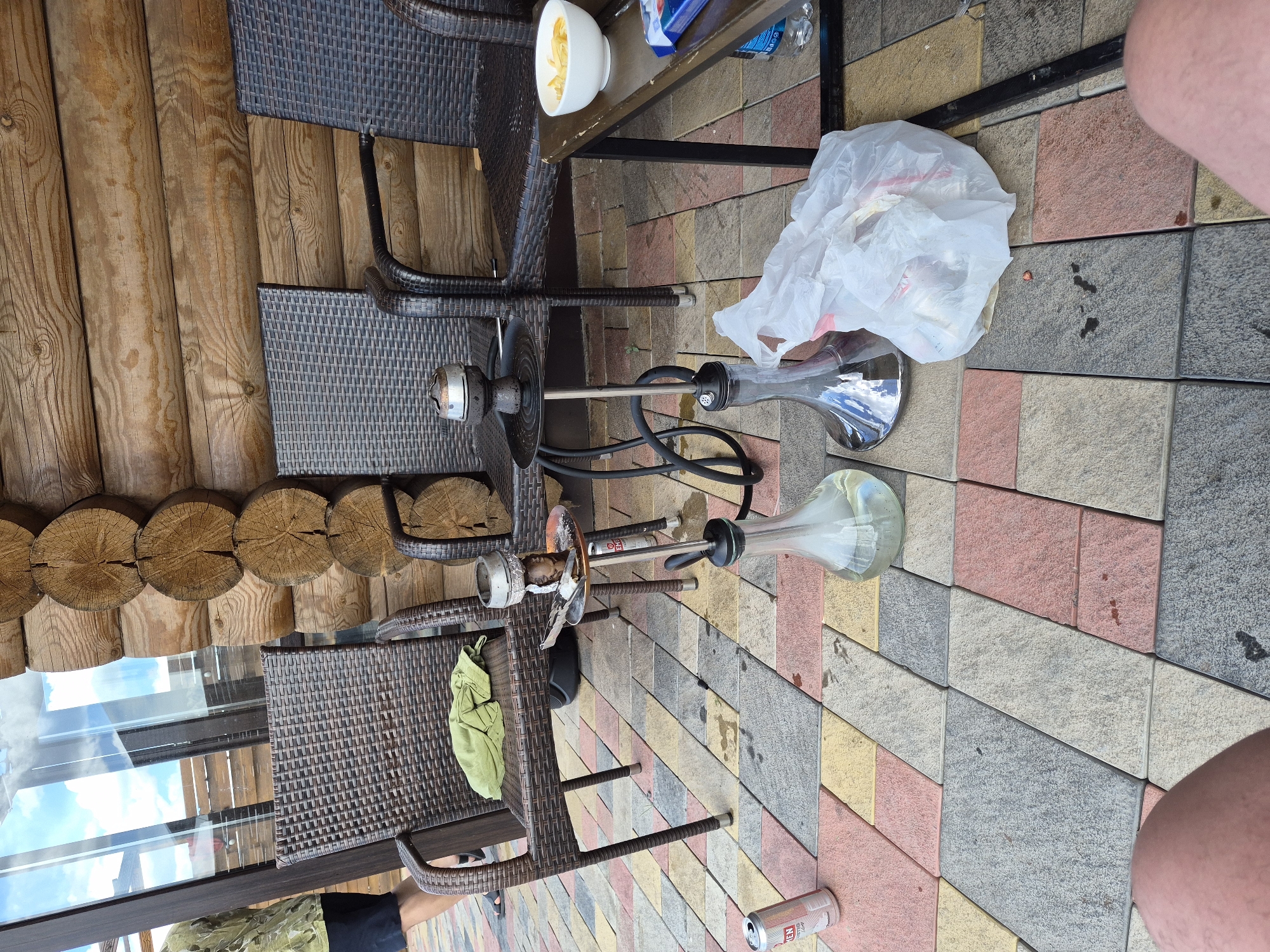The image depicts a ground-level outdoor setting, likely a patio or veranda, adjacent to a wooden log cabin structure. The ground is paved with interlocking rectangular tiles in various shades of grey, pink, and yellow. In the foreground, two human legs are visible, suggesting a person is seated or lying down.
Several objects are arranged around the area. Two wicker chairs are placed on the tiled ground. On one chair, a crumpled green cloth is visible. In front of the chairs, two hookahs are set up. The hookahs are made of glass and metal, with black hoses. One hookah has a metal bowl on top. A silver can, possibly a beverage, is lying on the ground near the chairs.
A small, dark-colored metal table is positioned to the right of the hookahs. On this table, a white bowl containing what appears to be potato chips is placed. To the right of the table, a white plastic bag filled with items is situated on the tiled ground. A partial view of a person wearing a camouflage-patterned shirt and dark shorts is visible on the far left of the image, with their back to the camera. Reflections of the sky and a wooden structure are visible in the glass windows behind the chairs.
The context of Kosshy, Kazakhstan, is not directly deducible from the visual elements within the image itself. However, the presence of outdoor seating, hookahs, and a relaxed atmosphere suggests a recreational or hospitality setting. The wooden cabin architecture could be indicative of a dacha (summer house) or a similar type of recreational dwelling common in some regions. The sunny weather suggested by the bright lighting implies a daytime scene.