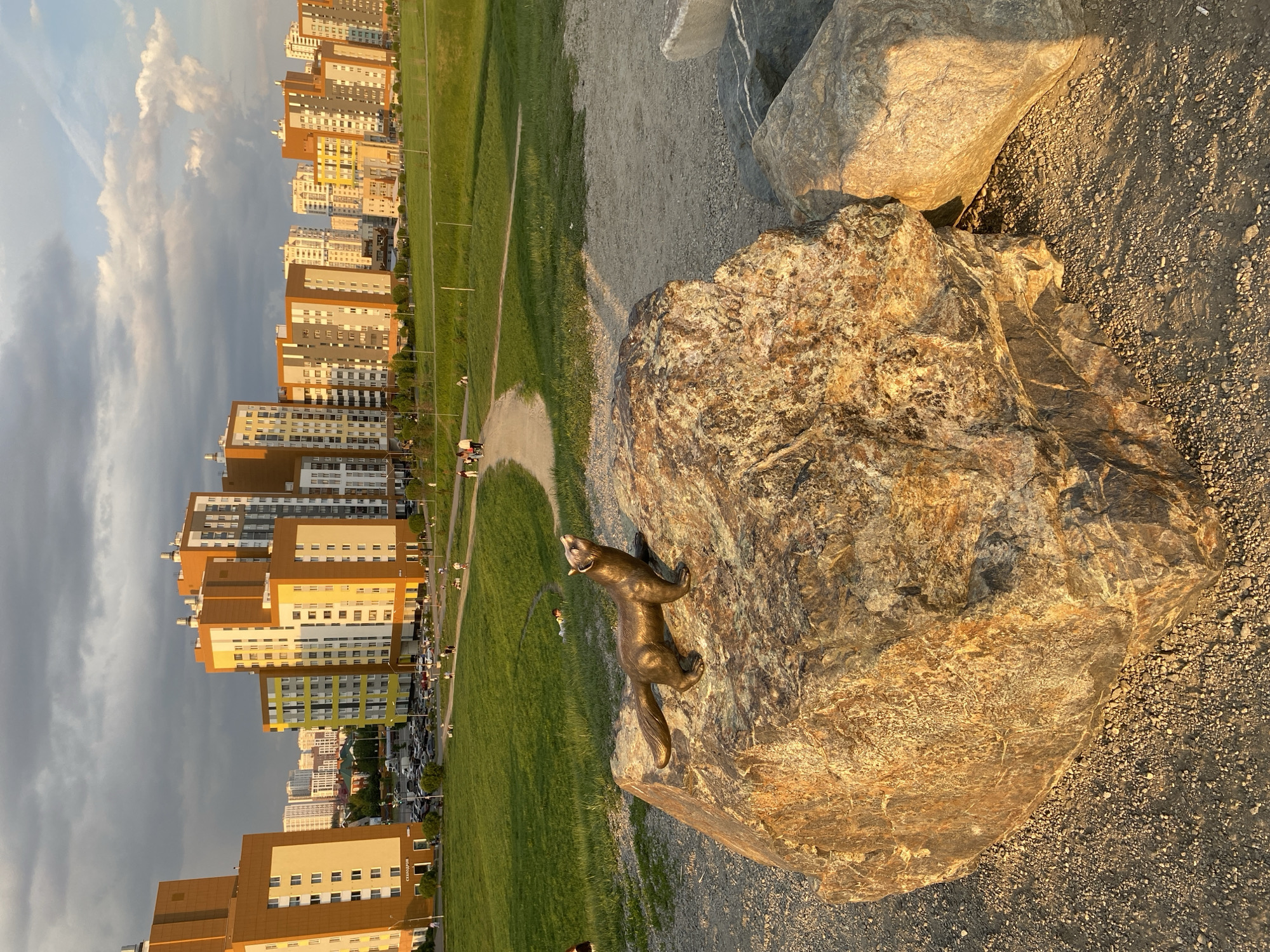 Here's a detailed description of the image:
**Scene:** A bronze statue of a sable is perched atop a large rock. In the background, there is a cityscape with tall, modern apartment buildings of various colors (beige, brown, yellow, green) and designs. A green park area with walking paths and scattered people stretches out between the rock and the buildings.
**Environment:** The weather is partly cloudy, with sunlight illuminating the rock and statue. The ground around the rock appears to be a gravel or pebble surface.
**Subject:** The statue of the sable is the focal point, standing out due to its metallic sheen and its placement on the rock. The surrounding rocks and buildings create a sense of scale and context.
**Location:** Based on the architectural style of the buildings and the presence of the sable statue (a local symbol), the location is likely within or near the city of Yekaterinburg, Russia.