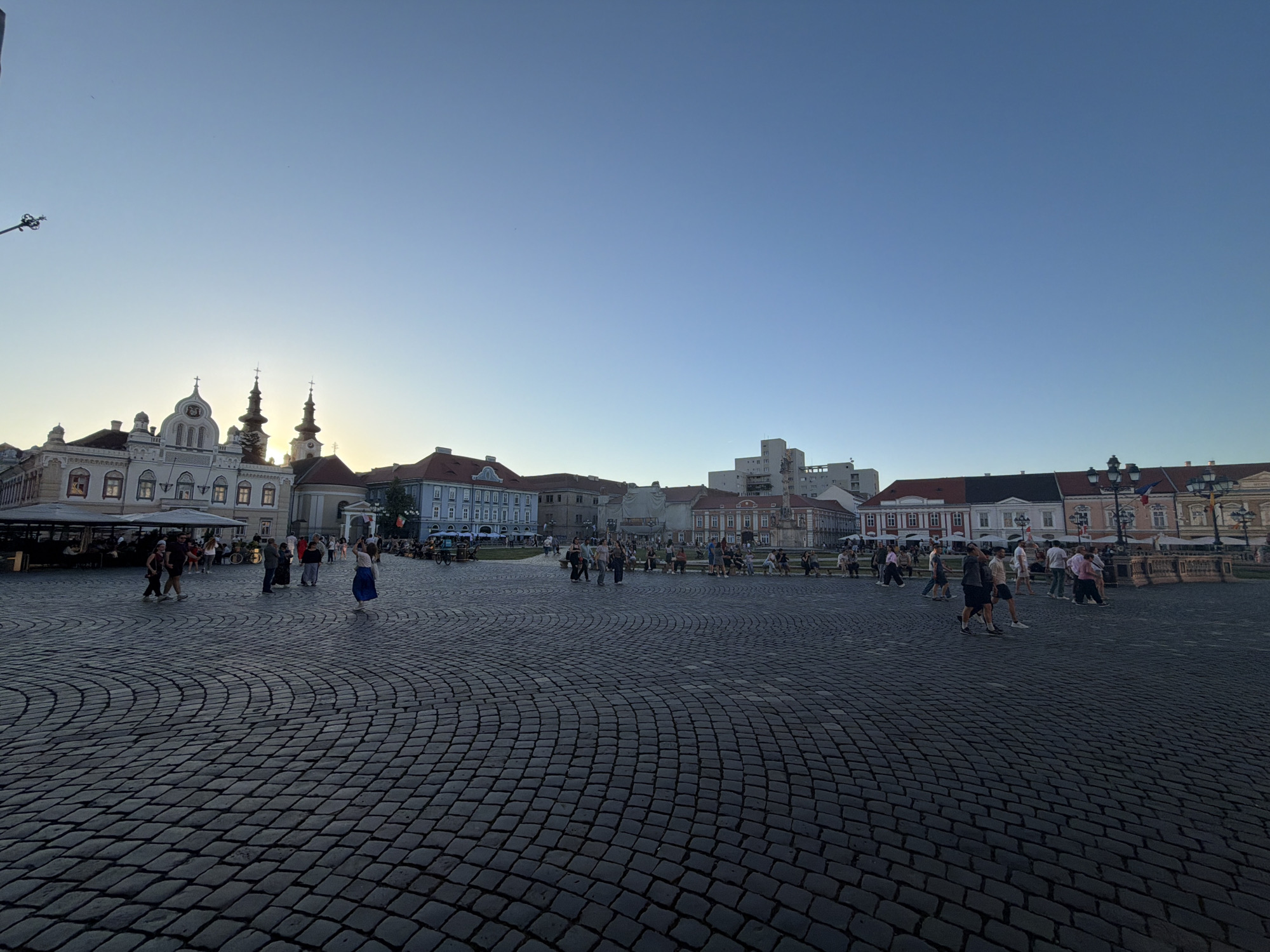 The image depicts a wide-angle view of Piața Unirii (Union Square) in Timișoara, Romania. The foreground is dominated by a cobblestone pavement laid in a curved pattern. Several people are walking across the square, including individuals and small groups. Buildings surround the square, representing a mix of architectural styles with a predominant presence of historical structures. The buildings are of varying heights and colors, some with ornate facades and architectural details. The sky is clear and transitioning between daylight and twilight. The sun is setting behind some of the buildings on the left, casting a soft light across the scene. The overall impression is of a bustling public square in the early evening.