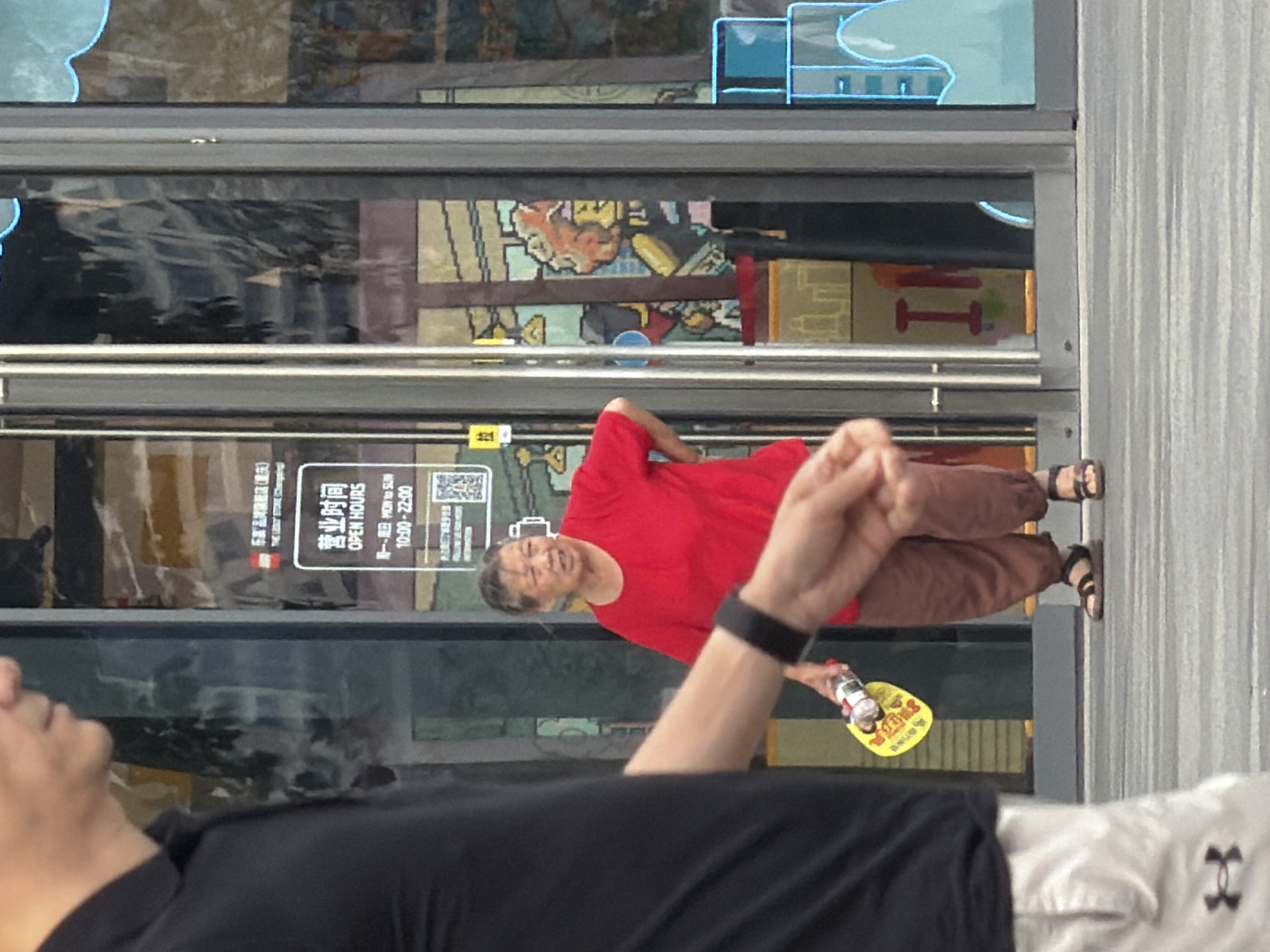 A woman in a red shirt and brown pants walks past a storefront in Yuzhong District, China. The woman is wearing sandals and carrying a yellow fan with a bottle. She is walking away from the camera. In the foreground, a person wearing a black shirt and white shorts is partially visible, with their arm extending into the frame, holding the fan and bottle. The storefront has glass doors with signage that includes "OPEN HOURS" and "10:00-22:00". The background appears to be a colorful display within the store. The ground is paved with gray and white stripes.