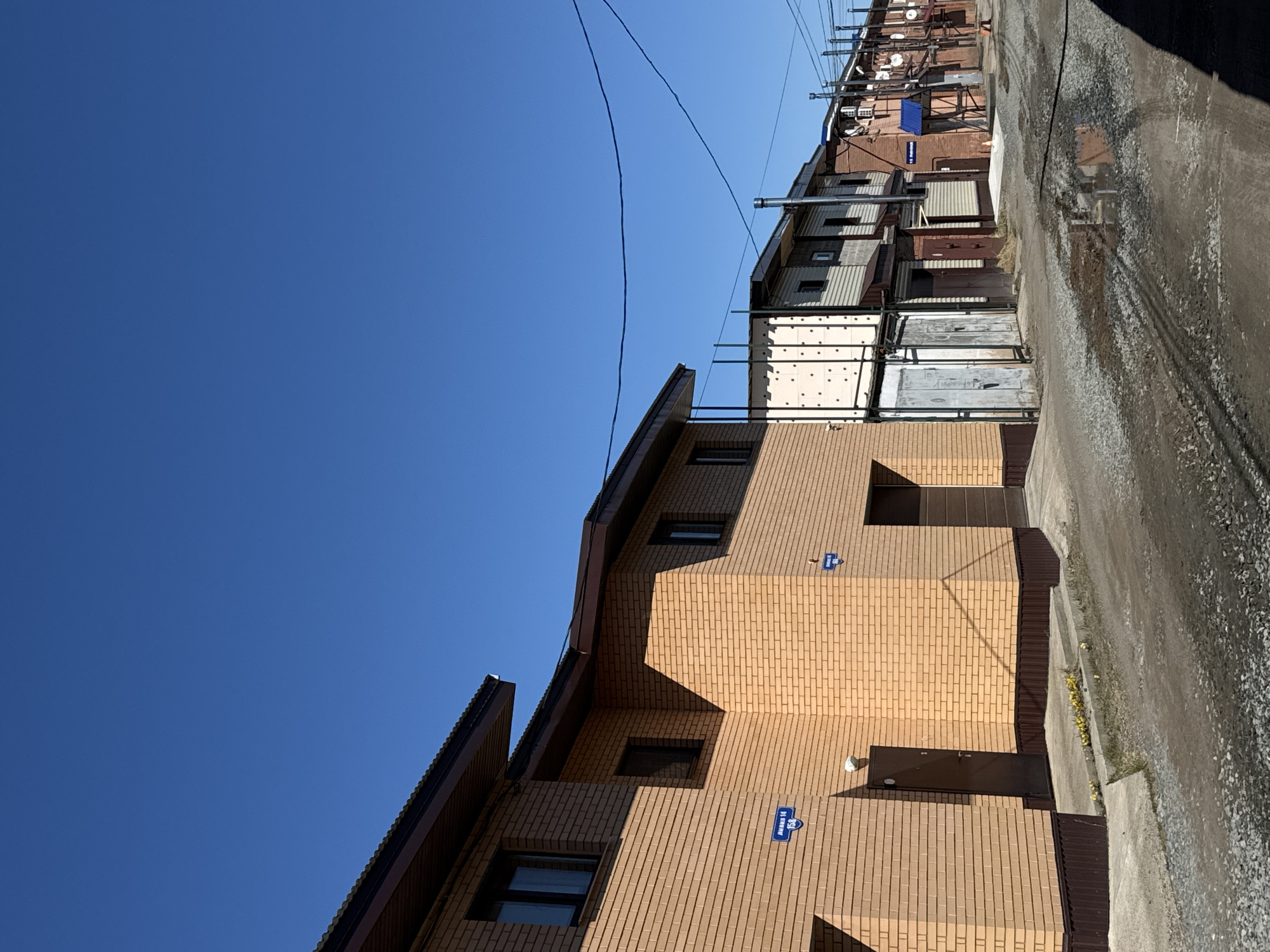 The image depicts a street scene in Chaikovsky, Russia. A multi-story brick building occupies the lower portion of the frame, its façade featuring several windows, two doors, and blue address plaques. The building's roofline is dark-colored. Above and to the right, a row of older, varied structures lines the street. These buildings are a mix of brick, siding, and metal, some with satellite dishes. A utility pole with wires runs across the top of the frame against a clear blue sky. The street is paved and appears wet and uneven. There is no visible human activity in the scene.