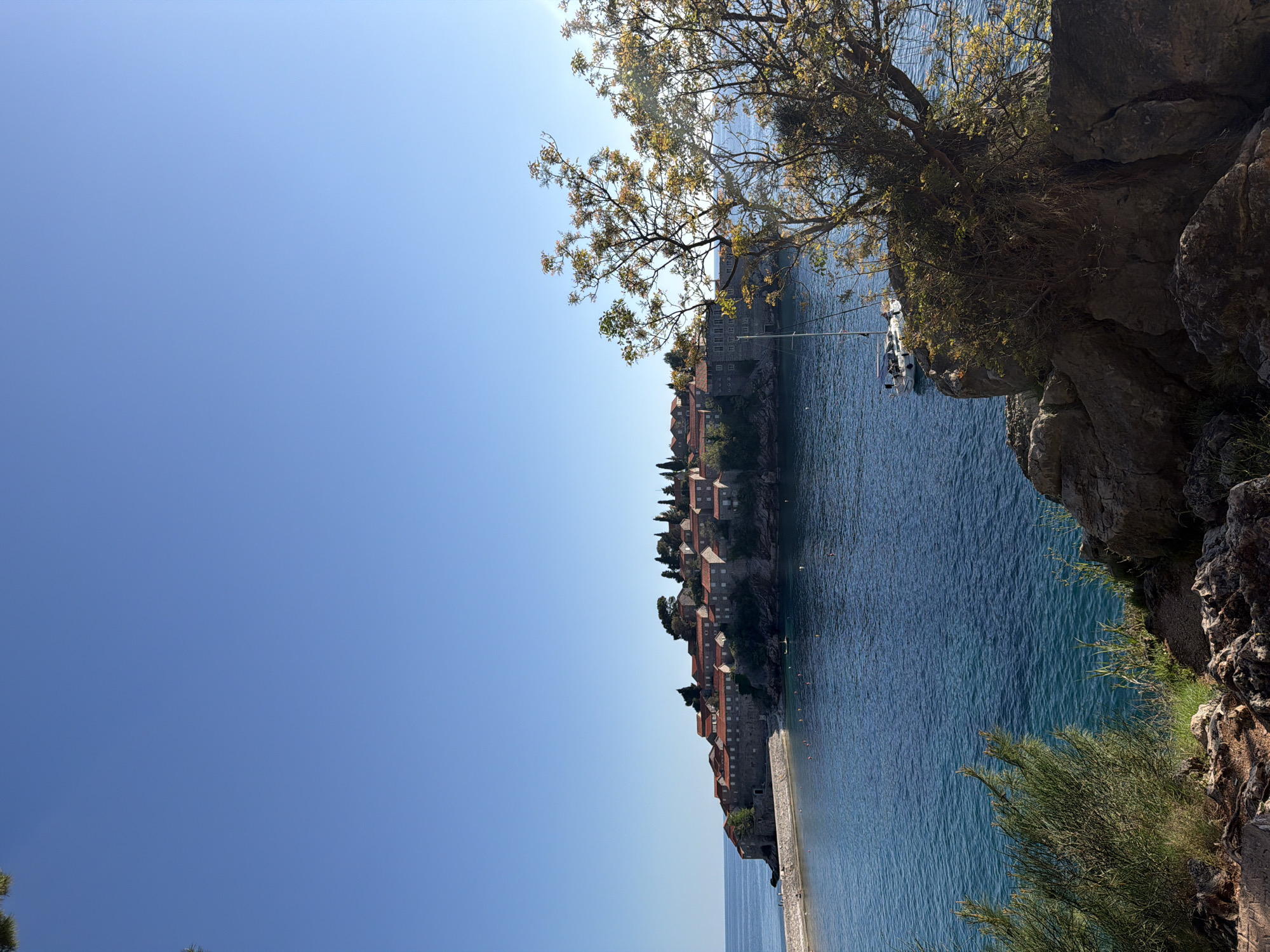 The image captures a view of Sveti Stefan, Montenegro, featuring the island-hotel in the center. The Adriatic Sea surrounds the island, with a sandy beach visible. Buildings with red-tiled roofs are constructed on the island. Vegetation, including trees and shrubs, appears along the rocky cliffside in the foreground. The sky is clear and blue. A small boat is moored near the island.