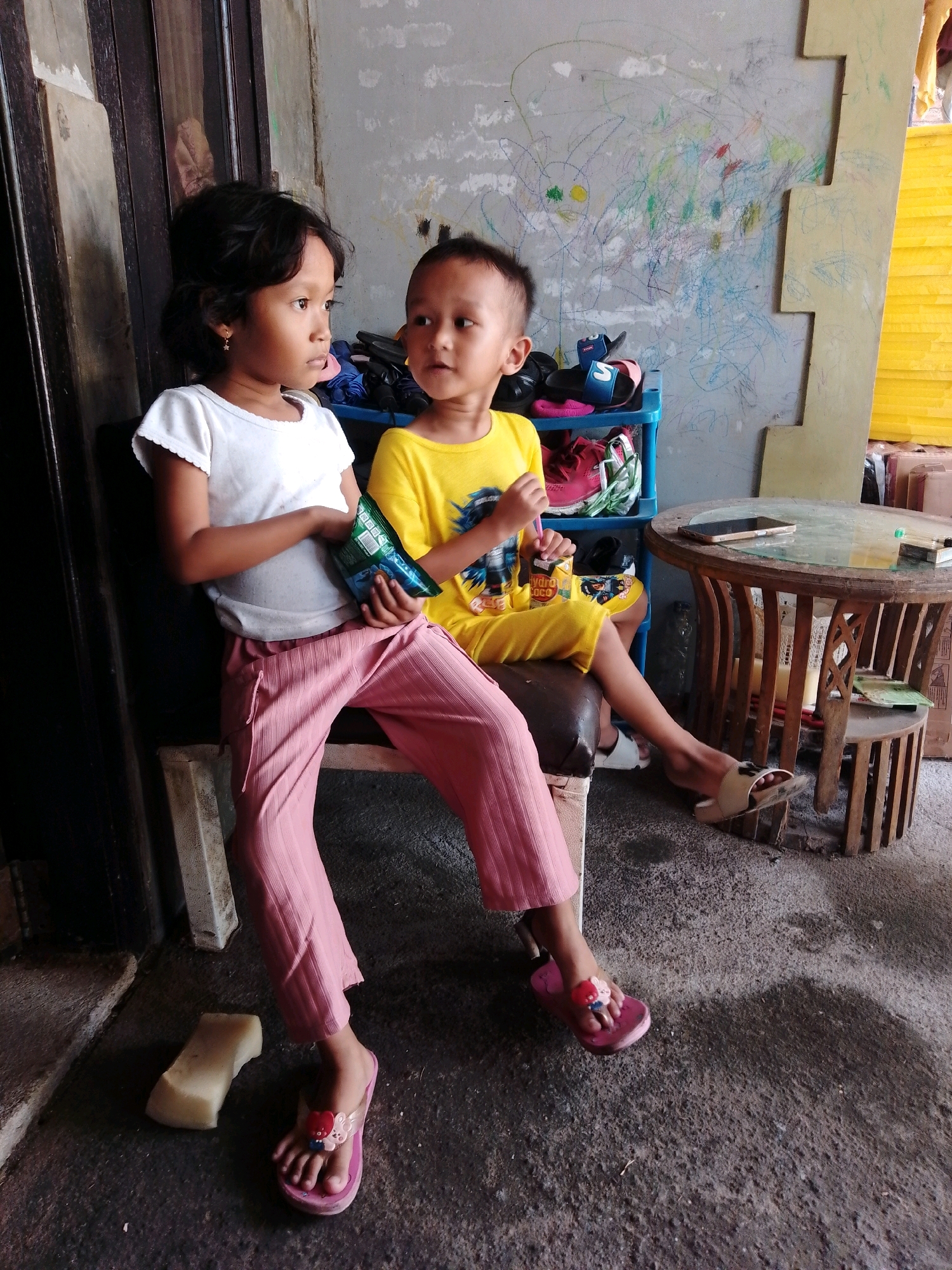 Two young children, a girl and a boy, are seated on a brown bench on a paved outdoor area. The girl, on the left, wears a white t-shirt and pink striped pants. She is holding a green snack bag. The boy, on the right, wears a yellow t-shirt with a graphic print and yellow shorts. He is also holding a snack bag. Behind them, a blue plastic shelf unit holds various items, including shoes and bags. A wall with colorful crayon drawings is visible behind the shelf. To the right, a round wooden table with a glass top sits on the pavement, with a mobile phone and a cigarette pack on it. A small wooden stool is next to the table. The ground is rough concrete, with some puddles of water. The scene appears to be in an outdoor common area or alley in Cikal, Indonesia, with a rustic and slightly cluttered environment.