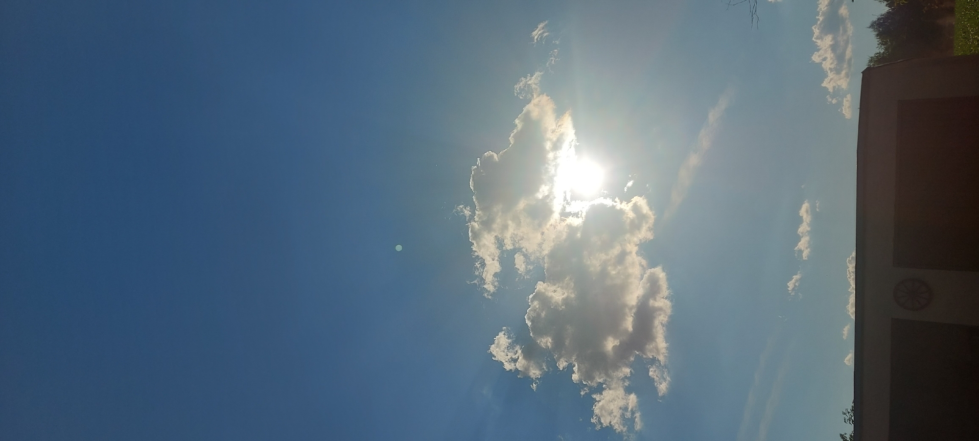This media file depicts a bright, sunny sky with scattered clouds. The sun is prominently visible, shining intensely and partially obscured by a large, fluffy cloud formation. The sky is a vibrant blue, suggesting daytime. In the lower right portion of the image, the edge of a building is visible. The building appears to be a light brown or beige color and has a dark rectangular section that could be a window or door. On the upper part of this building's facade, there's a circular decorative element that resembles a wagon wheel. In the distance, above the building, a line of green trees can be seen, indicating a natural landscape. The overall impression is one of a clear, pleasant day, likely late morning or early afternoon given the sun's position. There is no text visible in the image.