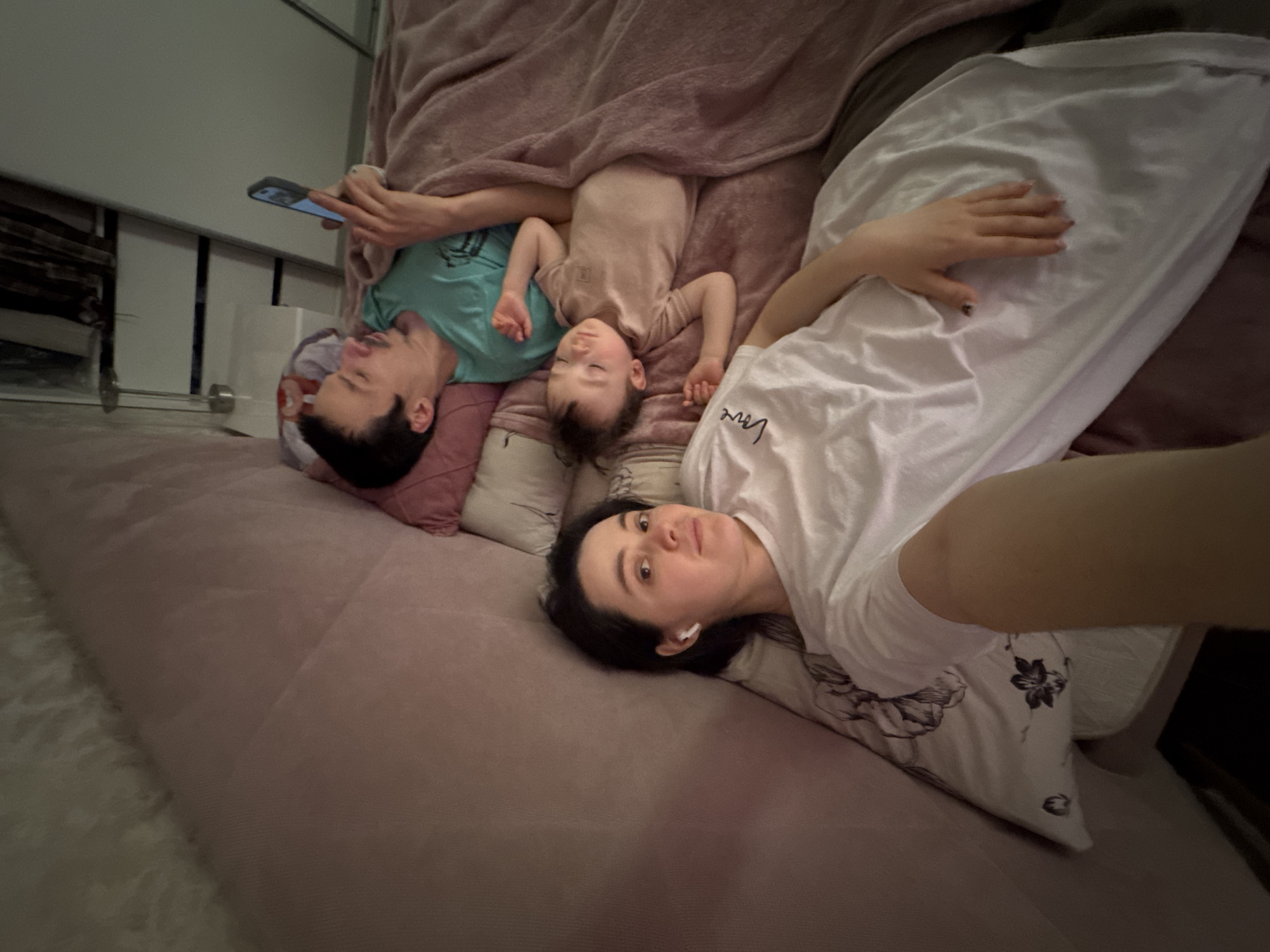 A medium-shot indoor image shows three individuals lying on a pink bed covered with a pink blanket.
A woman with dark hair and fair skin is lying on a pillow in the foreground, angled to the camera. She is wearing a white t-shirt with a floral design, and has a white wireless earbud in her left ear. Her right arm is partially visible, extending toward the camera.
Further back, a man with dark hair is lying on his back with a phone in his hand. He is wearing a light blue t-shirt and is resting on a red and black pillow.
A baby, dressed in light peach clothing, is lying between the man and the woman. The baby appears to be asleep.
The background shows a white storage unit with multiple compartments.
The location is not identifiable as Izhevsk, Russia, based on the visual information provided.