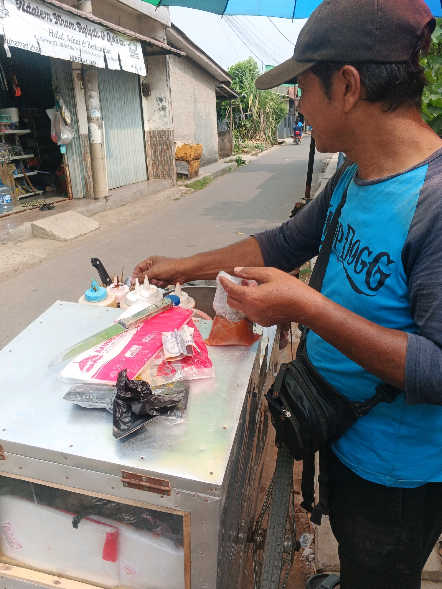 An Indonesian man wearing a blue t-shirt and a dark baseball cap stands behind a metal cart, which appears to be a food cart. He is in the process of preparing food, holding a small plastic bag with orange liquid in his right hand and reaching for a condiment bottle on the cart with his left. The cart is filled with various items, including plastic bags, a large knife, and multiple condiment bottles. In the background, a building with a prominent sign in Indonesian can be seen, which reads "Raidan Hj. Rham Raisyah & Qudwah Halal, Sehat & Berkhasiat". The cart is on a paved road, and there is a bicycle wheel visible to the right of the cart. The setting appears to be a street in the city of Kadu, Indonesia.