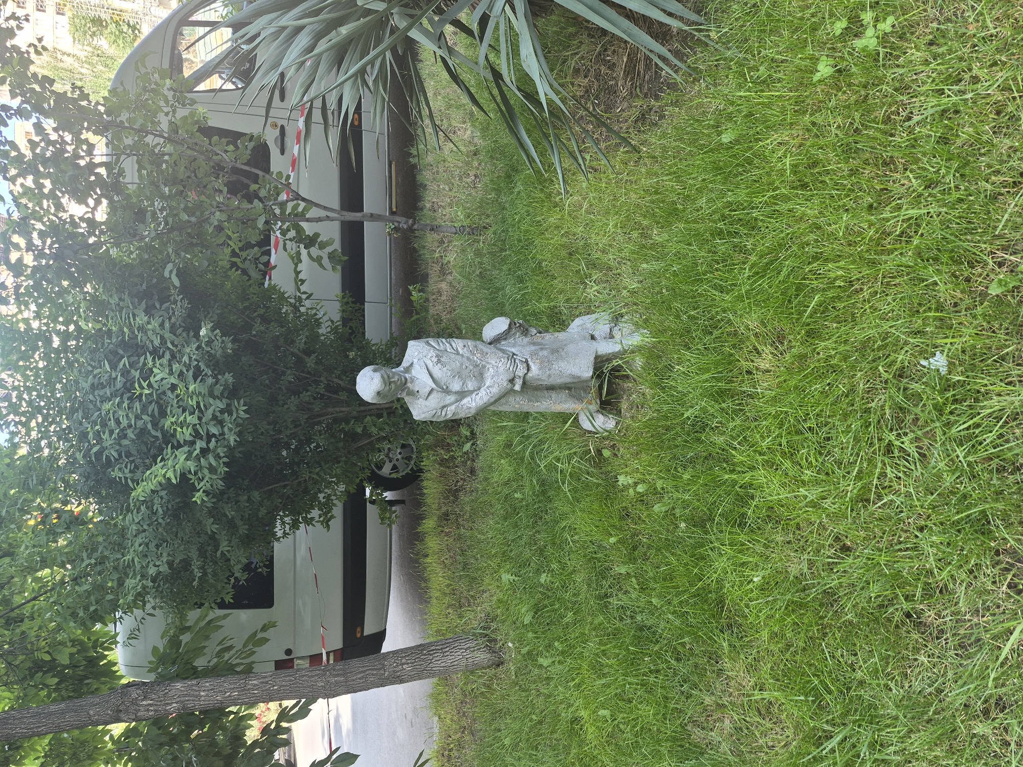 In Yerevan, Armenia, a small, weathered sculpture stands in a patch of green grass. The sculpture depicts two figures, one taller and standing, the other shorter and kneeling. Both figures are rendered in a muted, gray material, appearing to be made of stone or concrete. The standing figure wears a coat and appears to be looking down, while the kneeling figure is partially obscured.
Behind the sculpture, a white van is parked on a paved surface. The van has red and white striped tape draped across its side. Lush green foliage, including a tree with a textured bark and leafy branches, surrounds the scene. A spiky, palm-like plant is visible to the right of the sculpture. The grass is overgrown in some areas, with patches of dry leaves scattered on the ground. Sunlight filters through the leaves of the trees, creating dappled shadows on the grass.